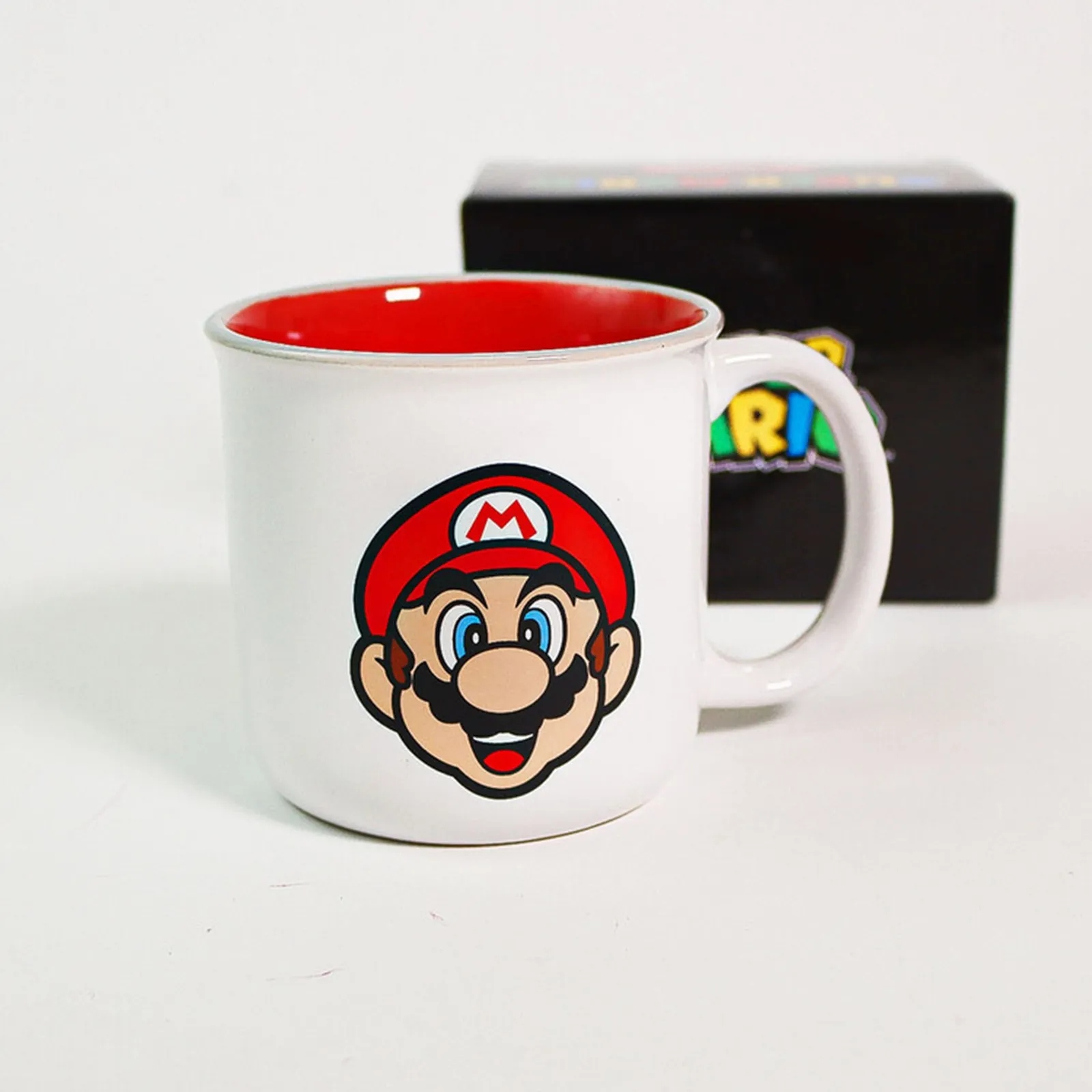 Una taza de cerámica blanca de Super Mario con interior rojo y caja negra al fondo sobre superficie clara.