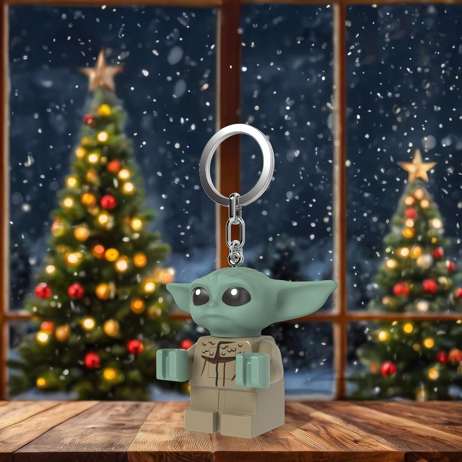 Un llavero de la figura de Yoda sobre una mesa de madera ante una ventana nevada con árboles de Navidad.