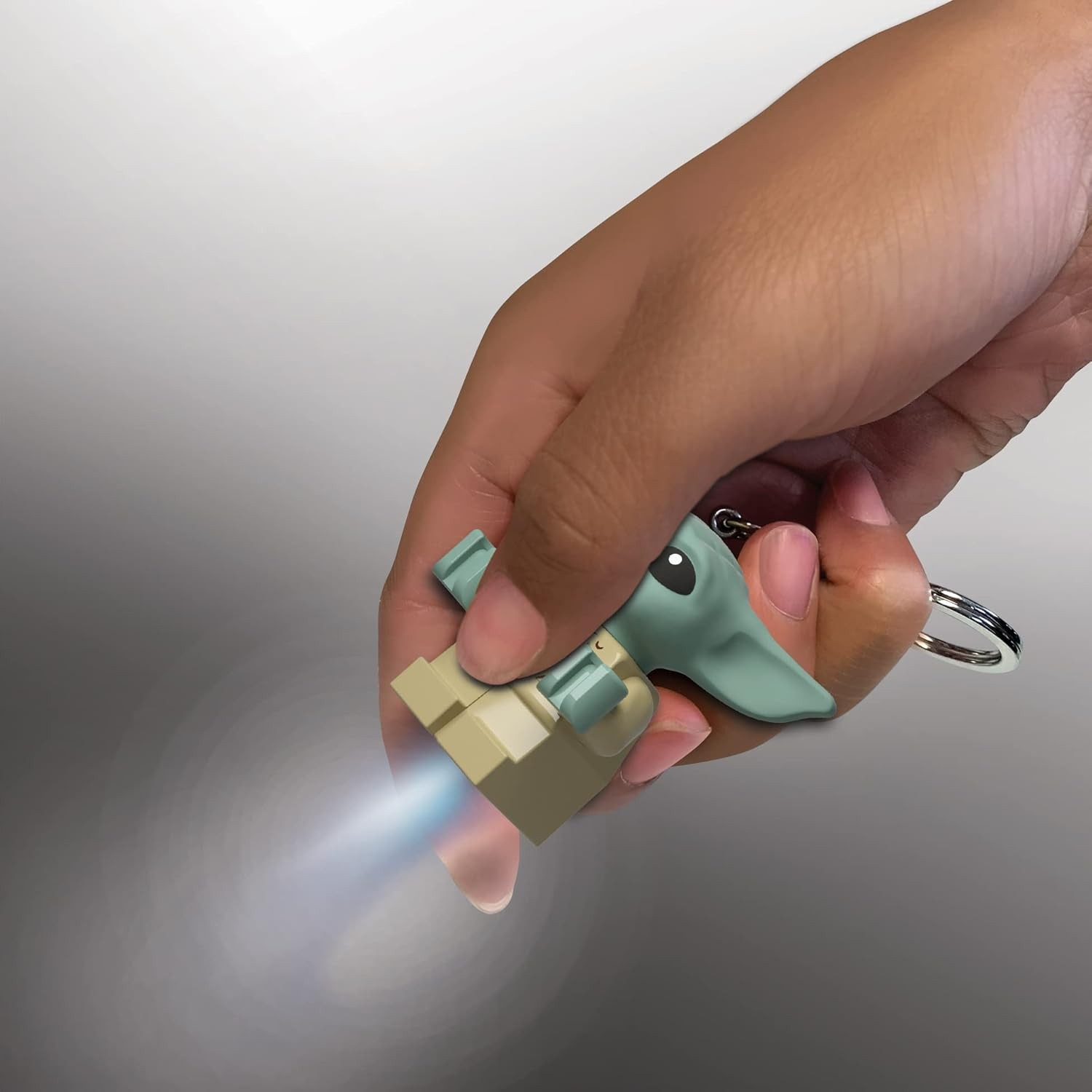 Una mano sostiene un llavero de la figura de Baby Yoda que emite una luz brillante desde su base sobre un fondo gris.
