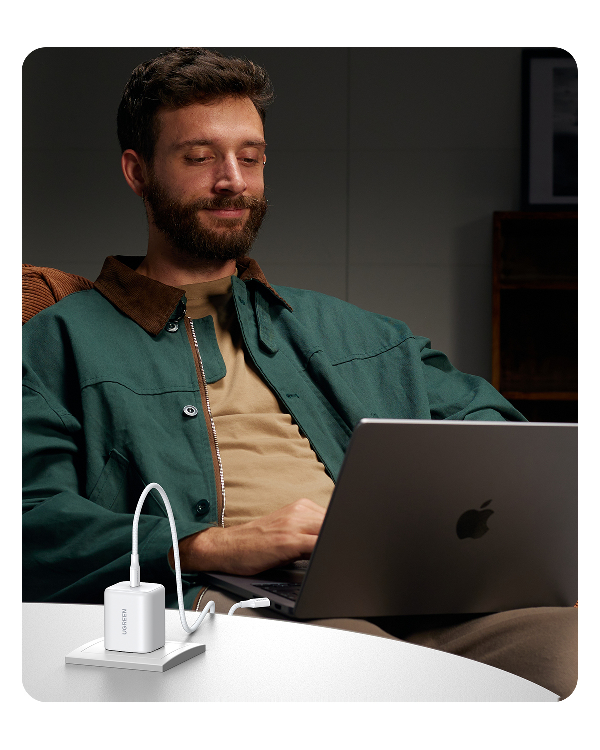 Sakalalı bir adam dizüstü bilgisayar kullanırken gülümsüyor, beyaz masada beyaz bir USB-C şarj cihazı takılı.