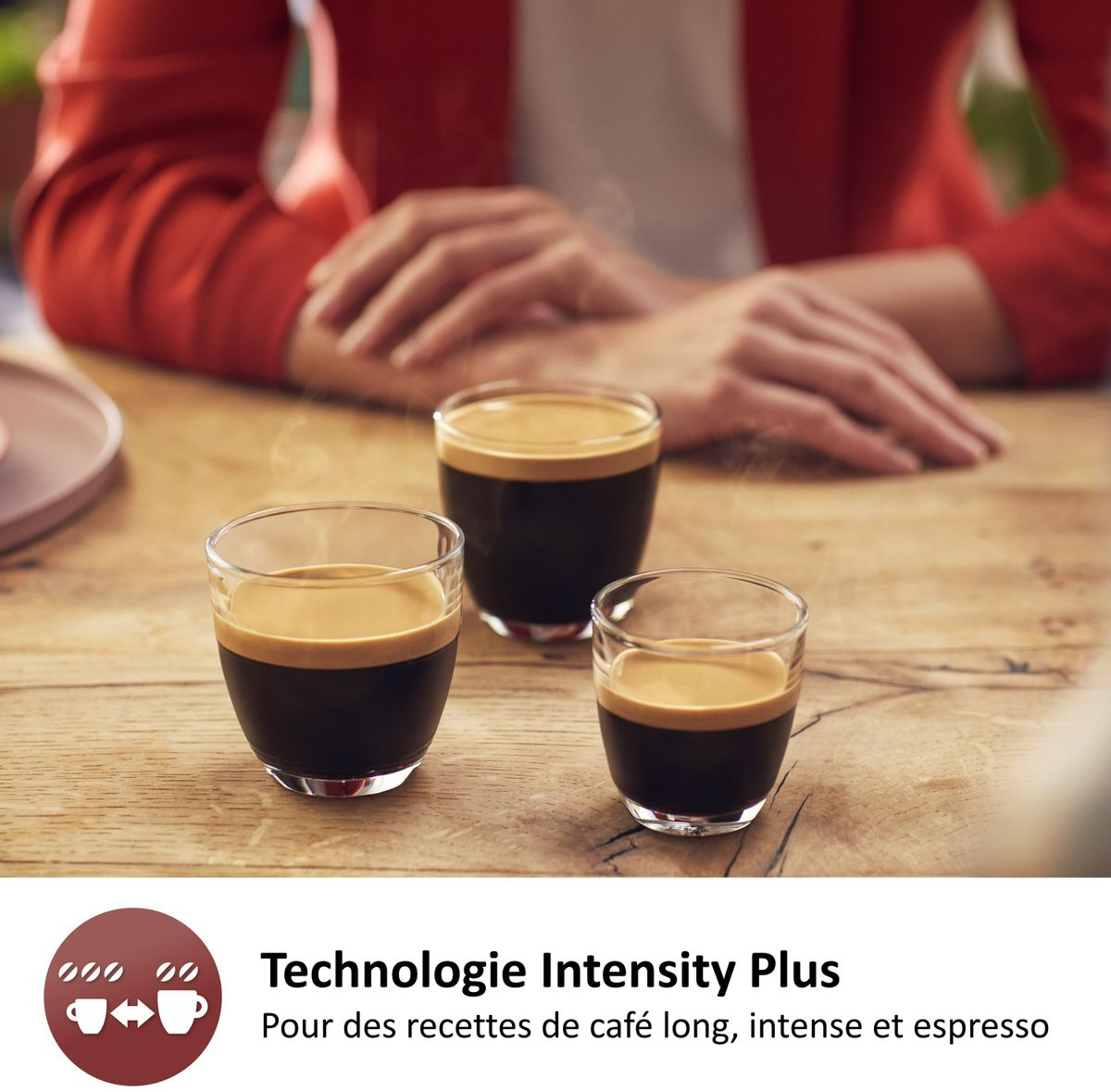 Drei kleine Gläser mit frisch gebrühtem Kaffee und Crema stehen auf einem Holztisch, im Hintergrund sitzt eine Person.