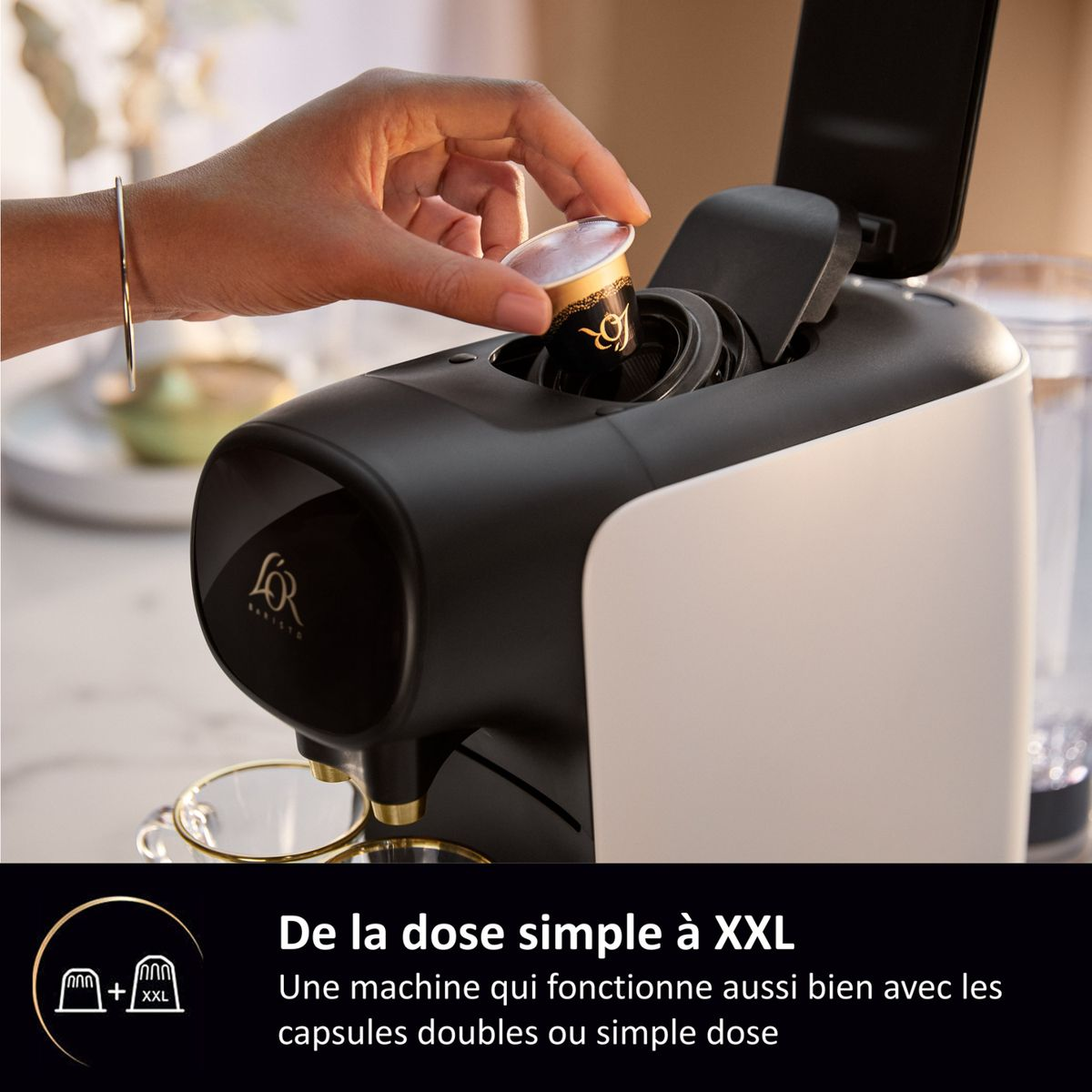 A hand inserts a capsule into a modern black and white coffee machine with glass cups below.