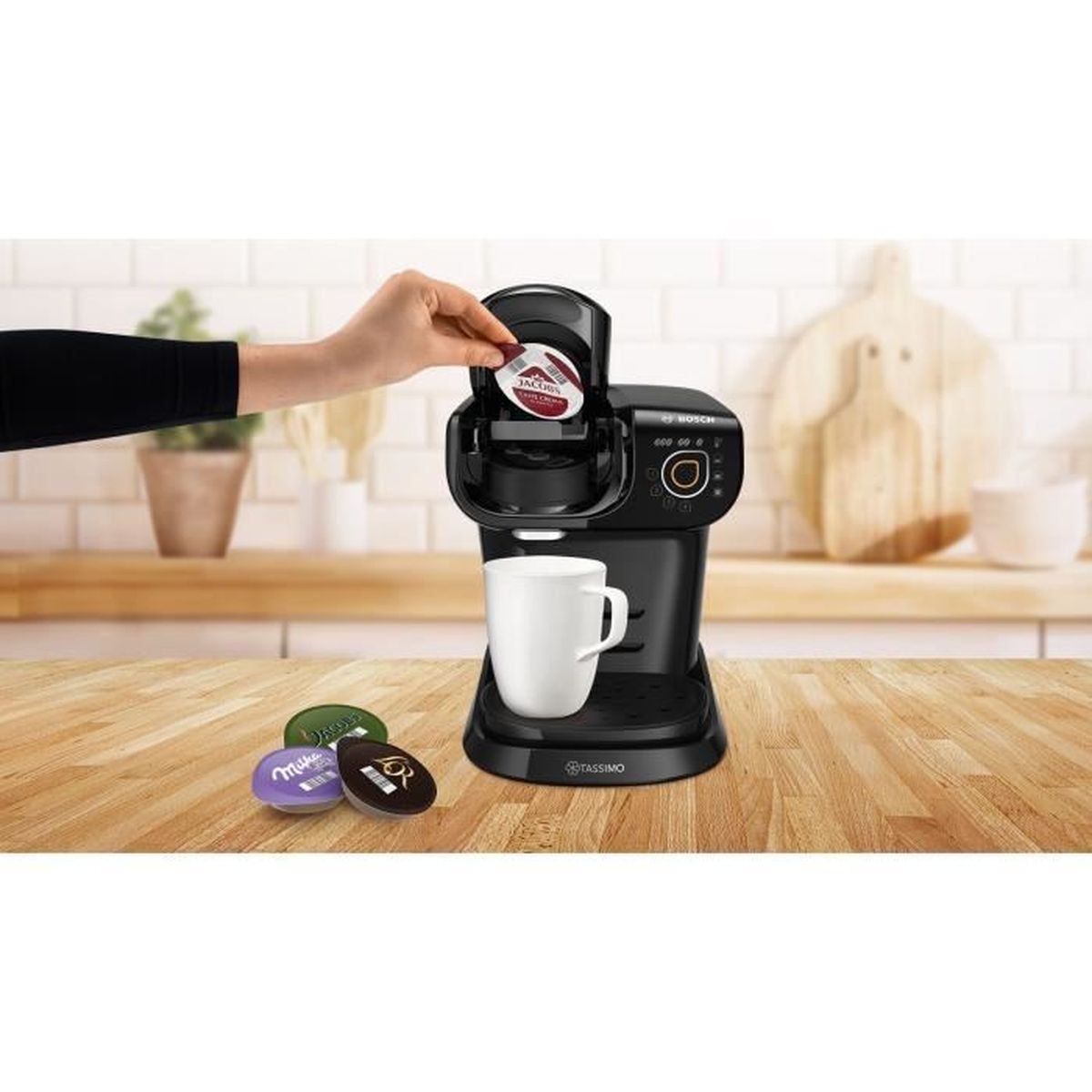 Una mano coloca una cápsula de café en una máquina Tassimo negra sobre una encimera de madera con taza blanca.