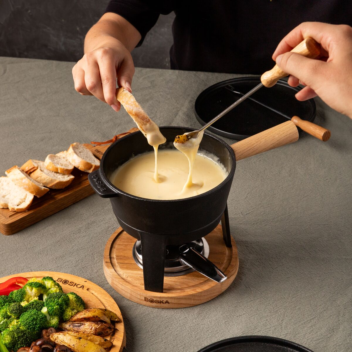 Personen tauchen Brot und Gemuese in einen Kasesuchetopf mit geschmolzenem Kase auf einem Holzstand.