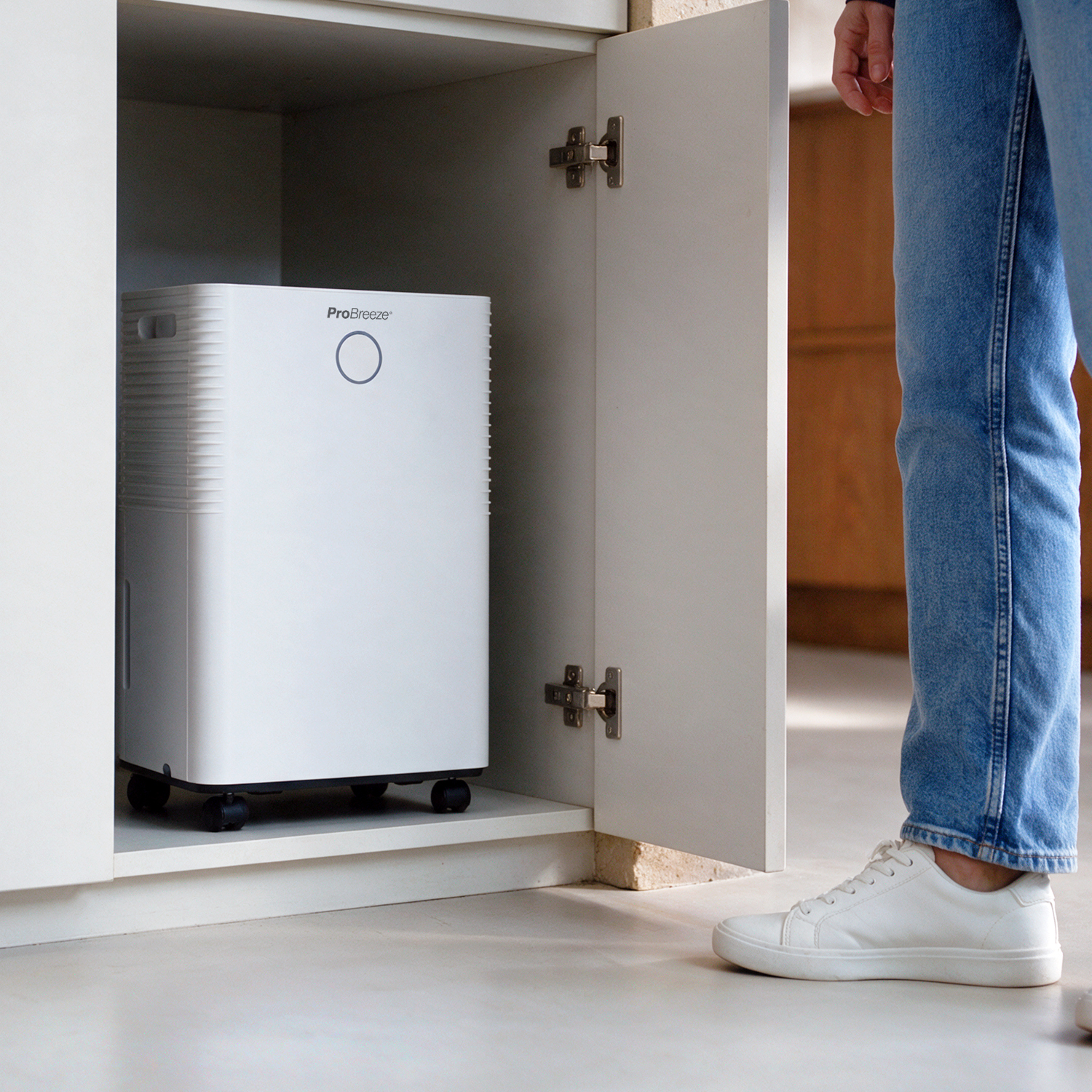 Ein weißer Pro Breeze Luftentfeuchter steht im offenen Unterschrank neben einer Person in Jeans und weißen Sneakers.
