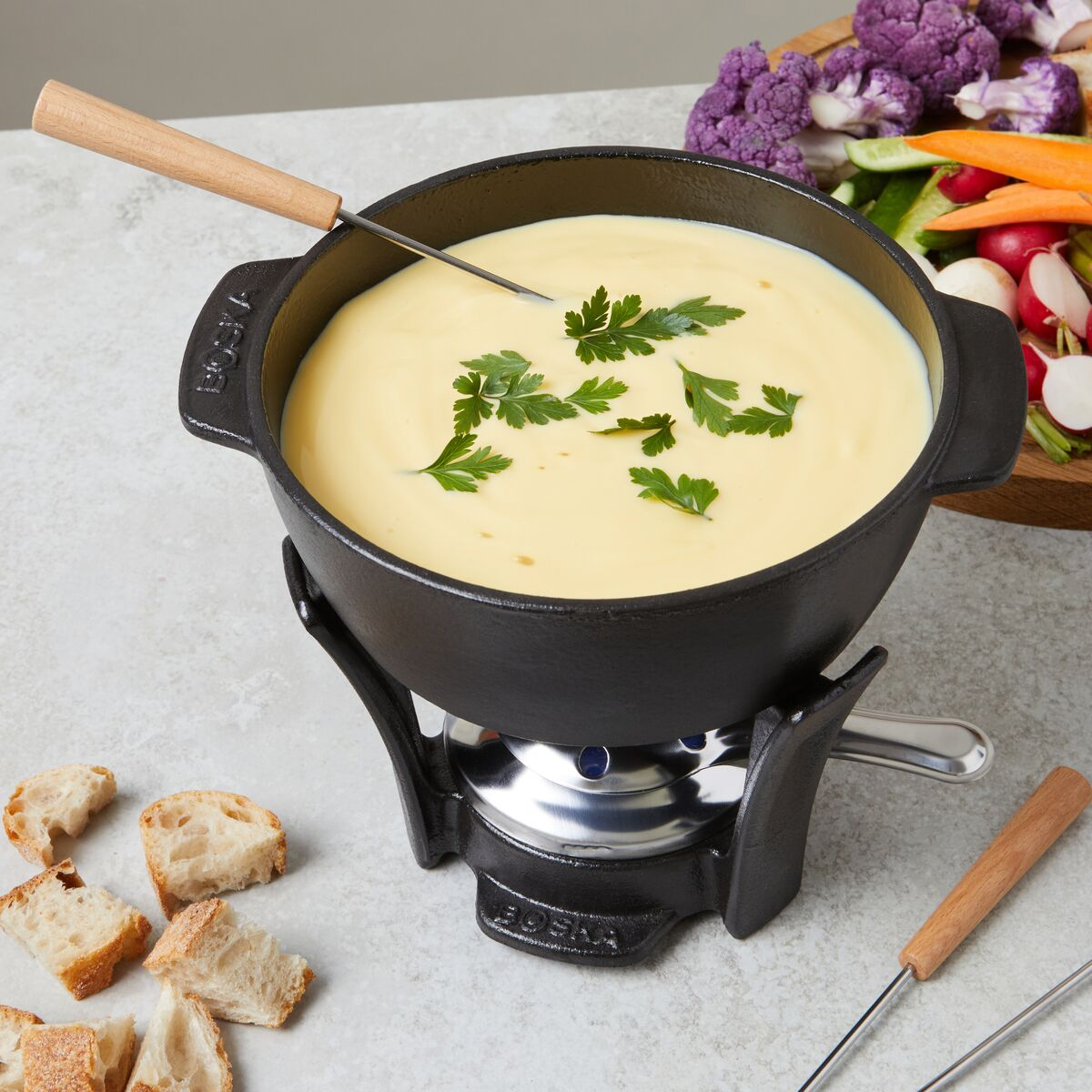 Ein schwarzer Fonduetopf mit geschmolzenem Käse und Petersilie, auf einem Brenner, mit Brotstücken und Gemüse.