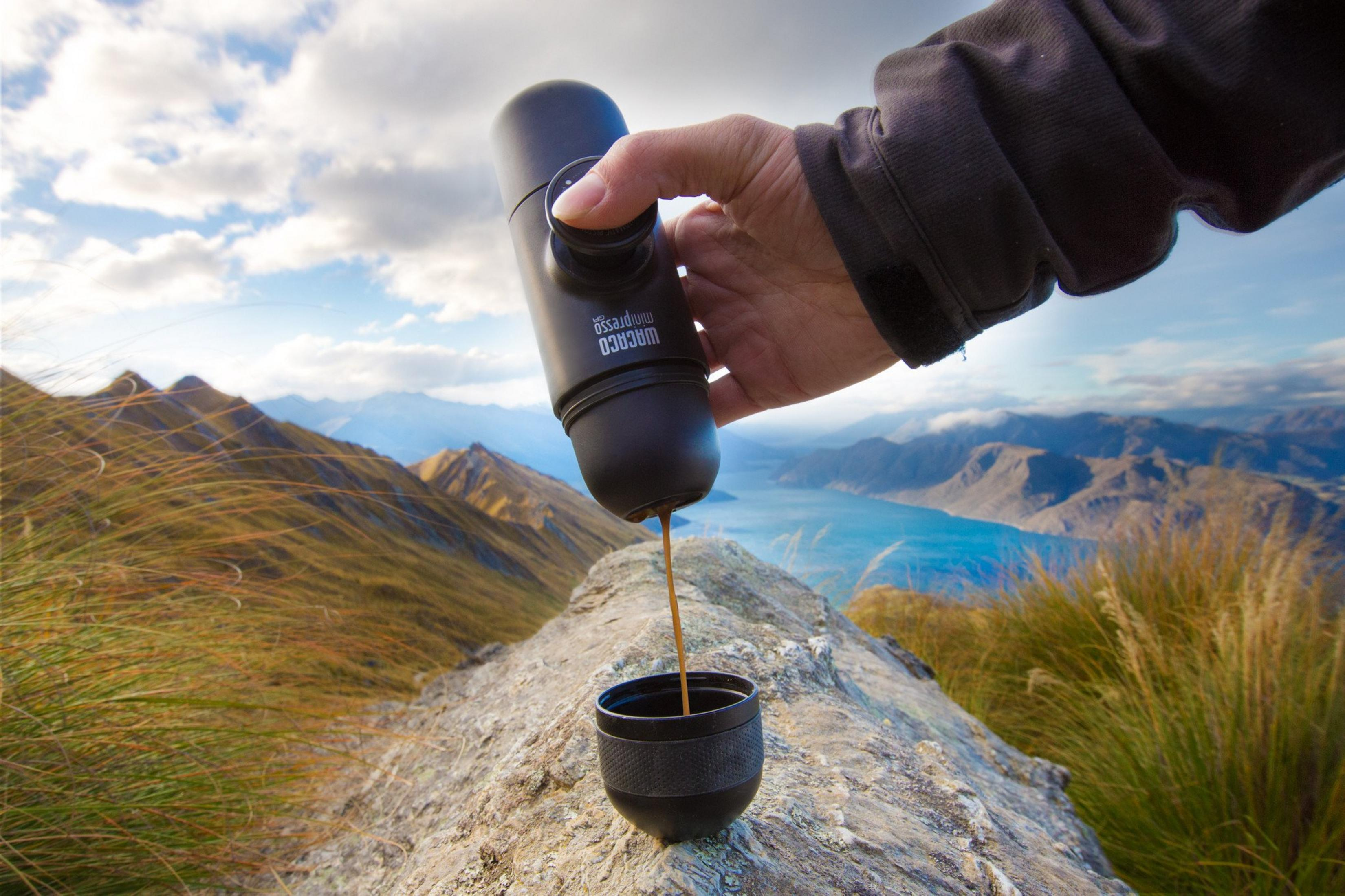 Une main pompe une cafetière portable sur une montagne, versant du café dans une tasse avec un lac en arrière-plan.