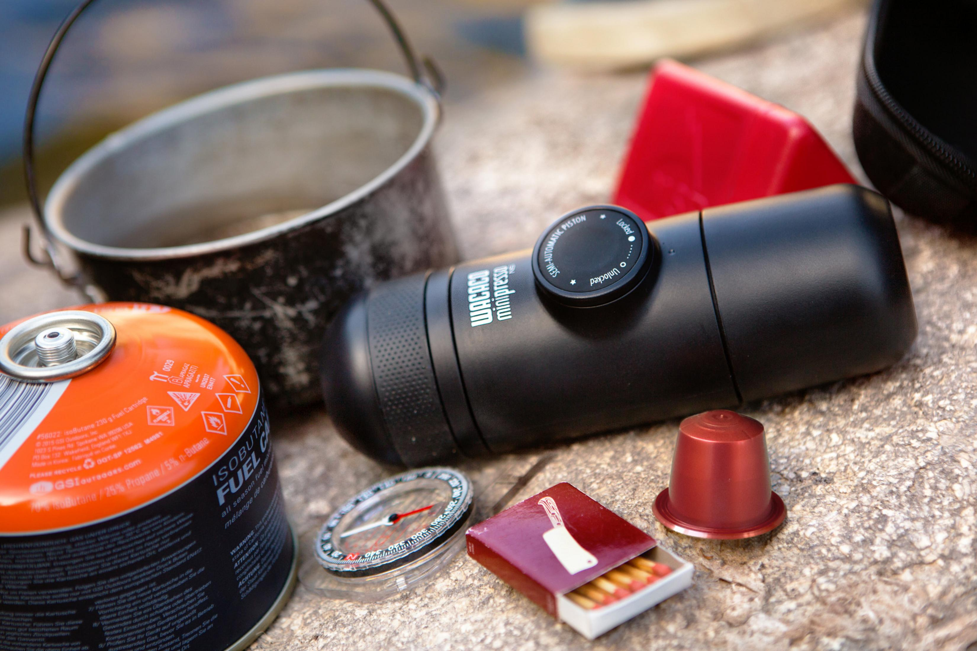 Casserole, cafetière manuelle noire, cartouche de gaz, boussole, allumettes et capsule de café sur pierre.