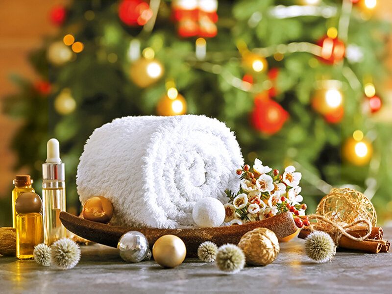 Escena de spa navideña con toalla blanca, aceites y bolas frente a un árbol.
