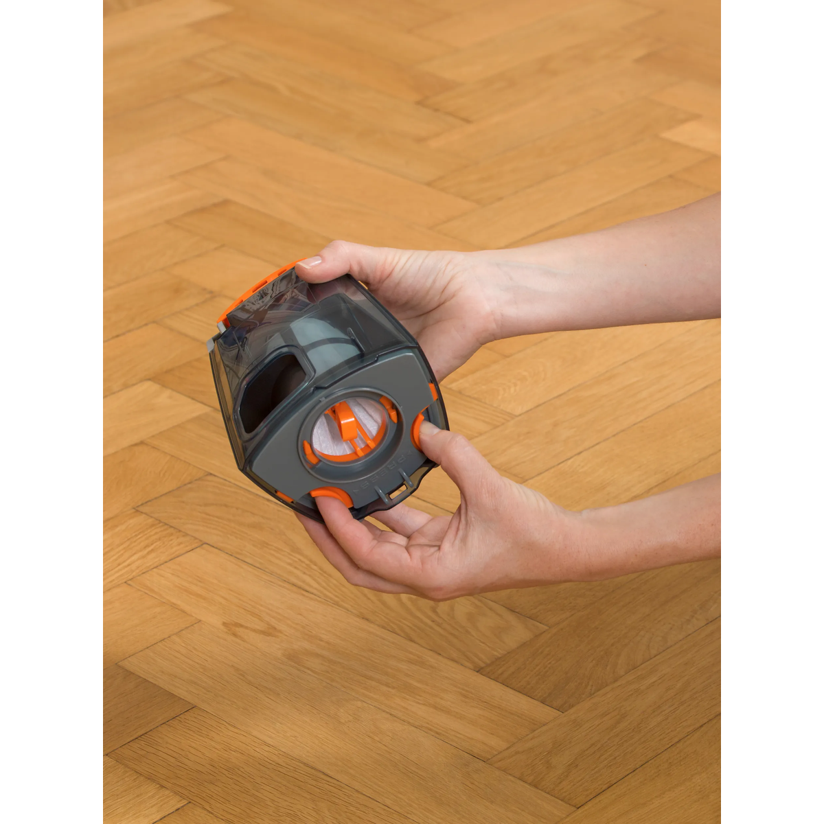 Manos sosteniendo un depósito de polvo de aspiradora gris y naranja sobre un suelo de madera.