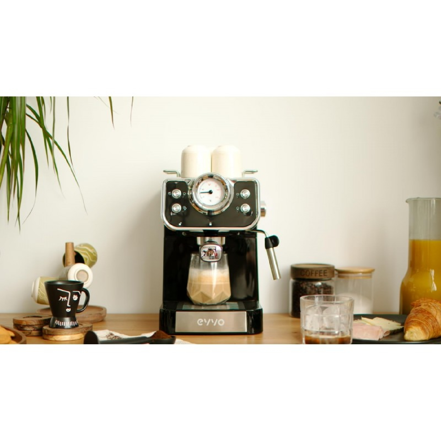 Cafetera espresso negra en encimera de madera, con tazas, vasos y un cruasán.