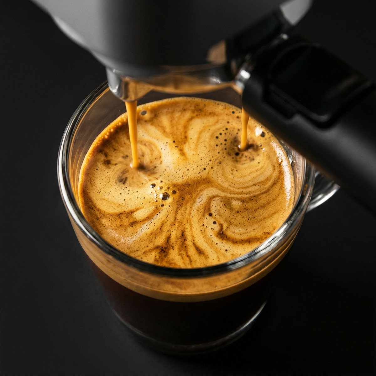 Un espresso fresco se vierte de una cafetera en una taza de vidrio transparente sobre fondo oscuro.