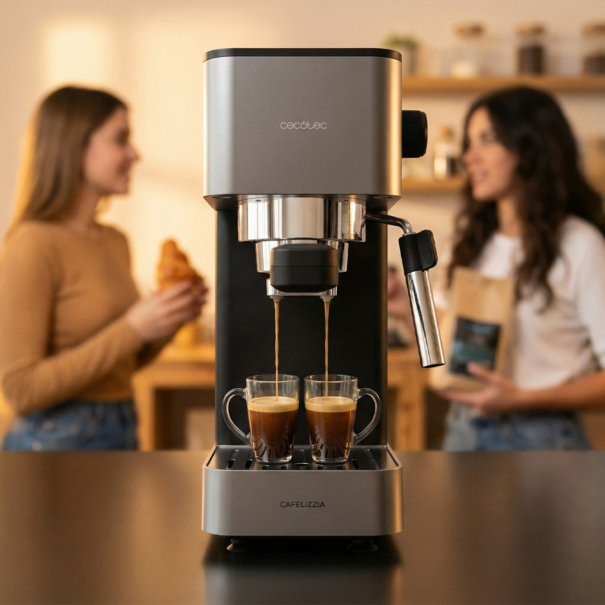 Una cafetera Cecotec prepara espresso en dos vasos. Al fondo, dos mujeres charlan en una cocina.