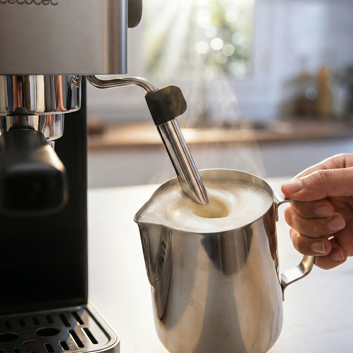 Un vaporizador espuma leche en una jarra de metal frente a una cafetera.