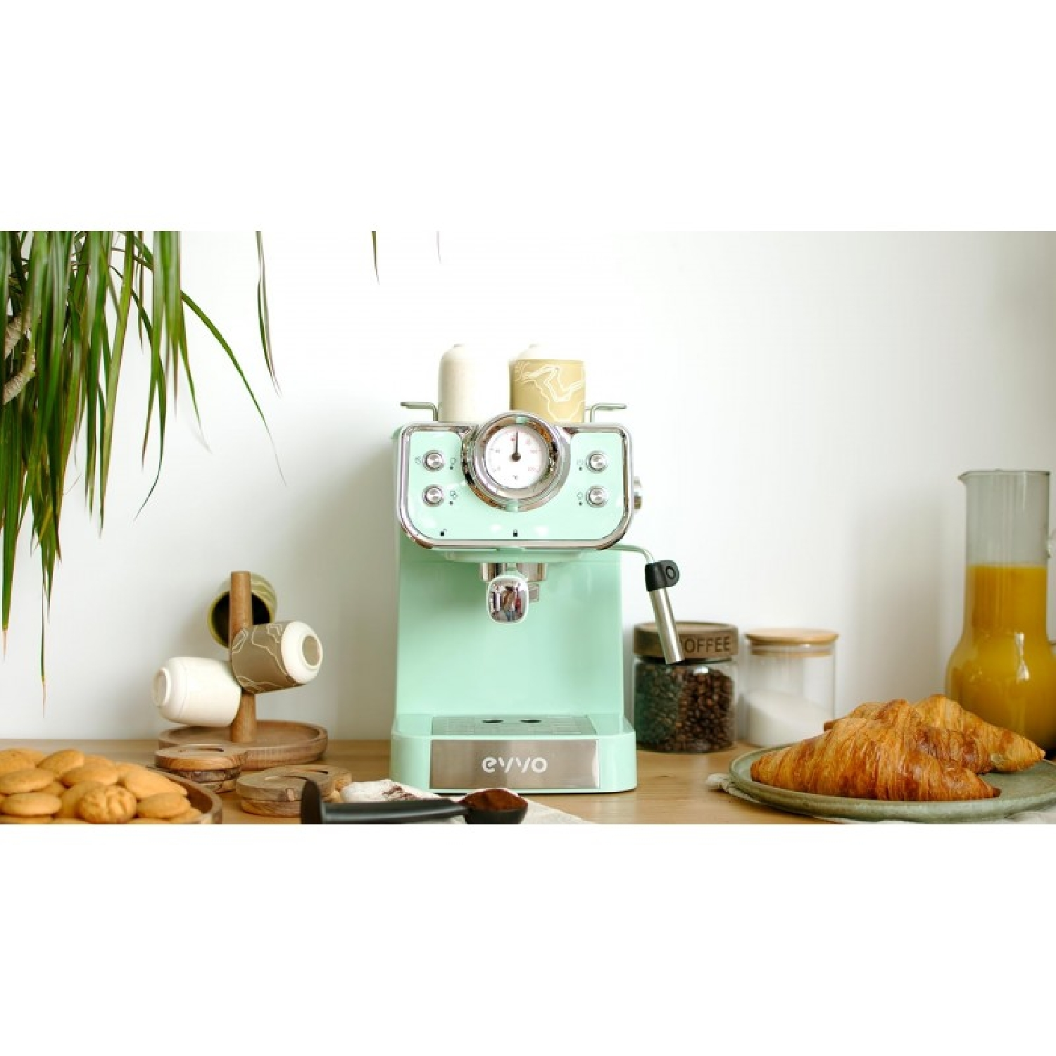 Cafetera espresso verde pastel en una mesa de madera, rodeada de cruasanes, galletas, granos de café y zumo de naranja.
