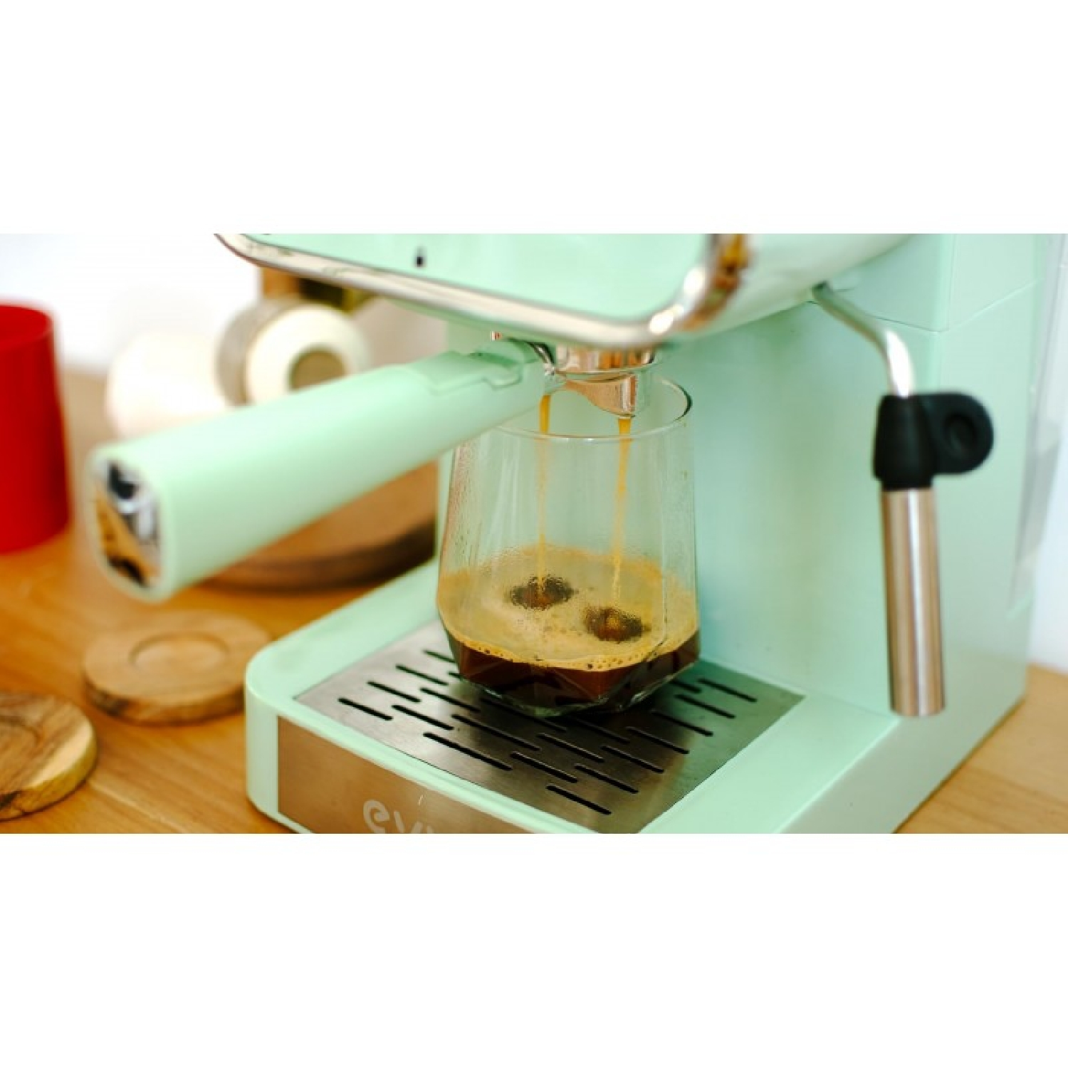 Una cafetera verde menta vierte café en un vaso sobre una barra de madera.