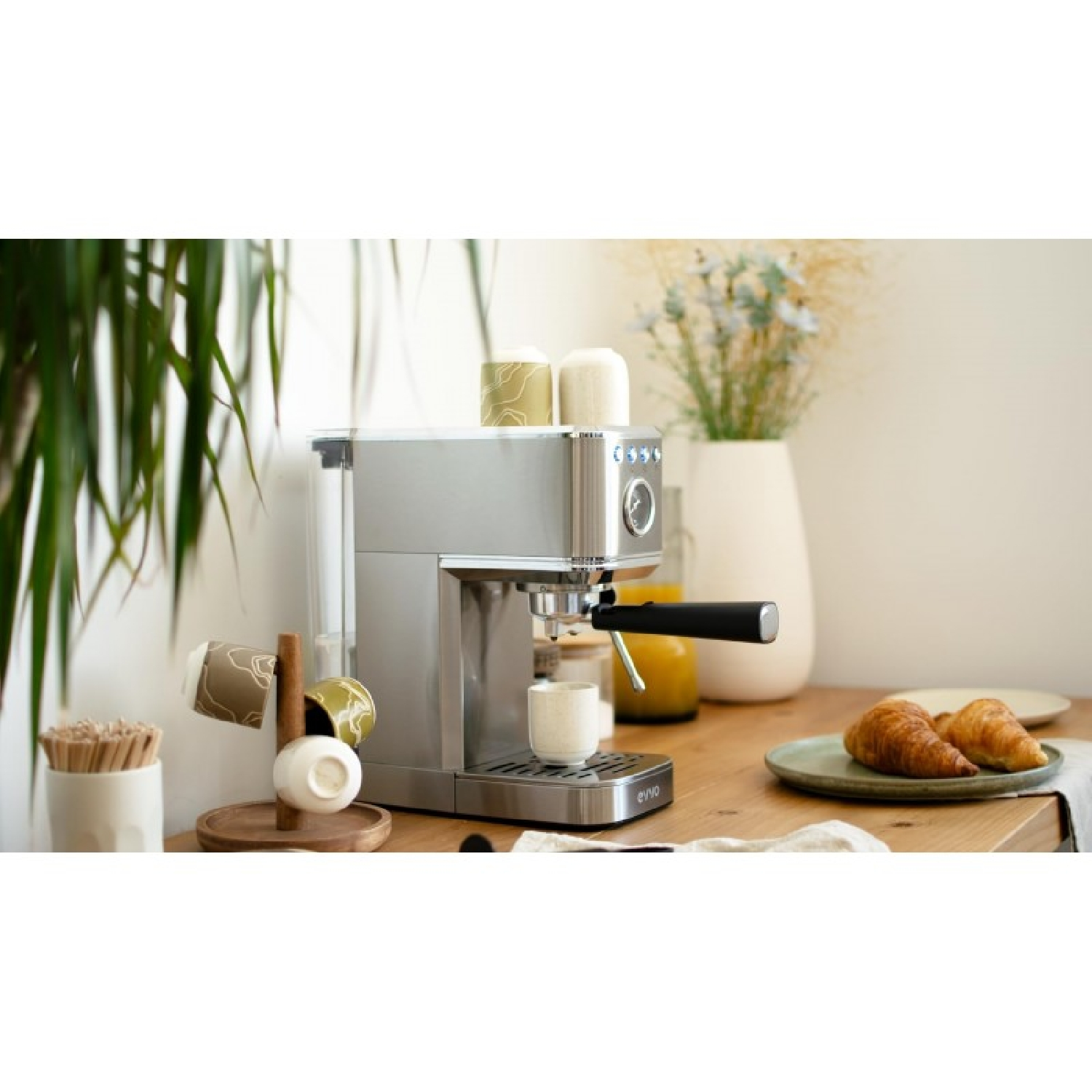 Una cafetera espresso plateada en una mesa de madera con croissants en un plato y un soporte para tazas.