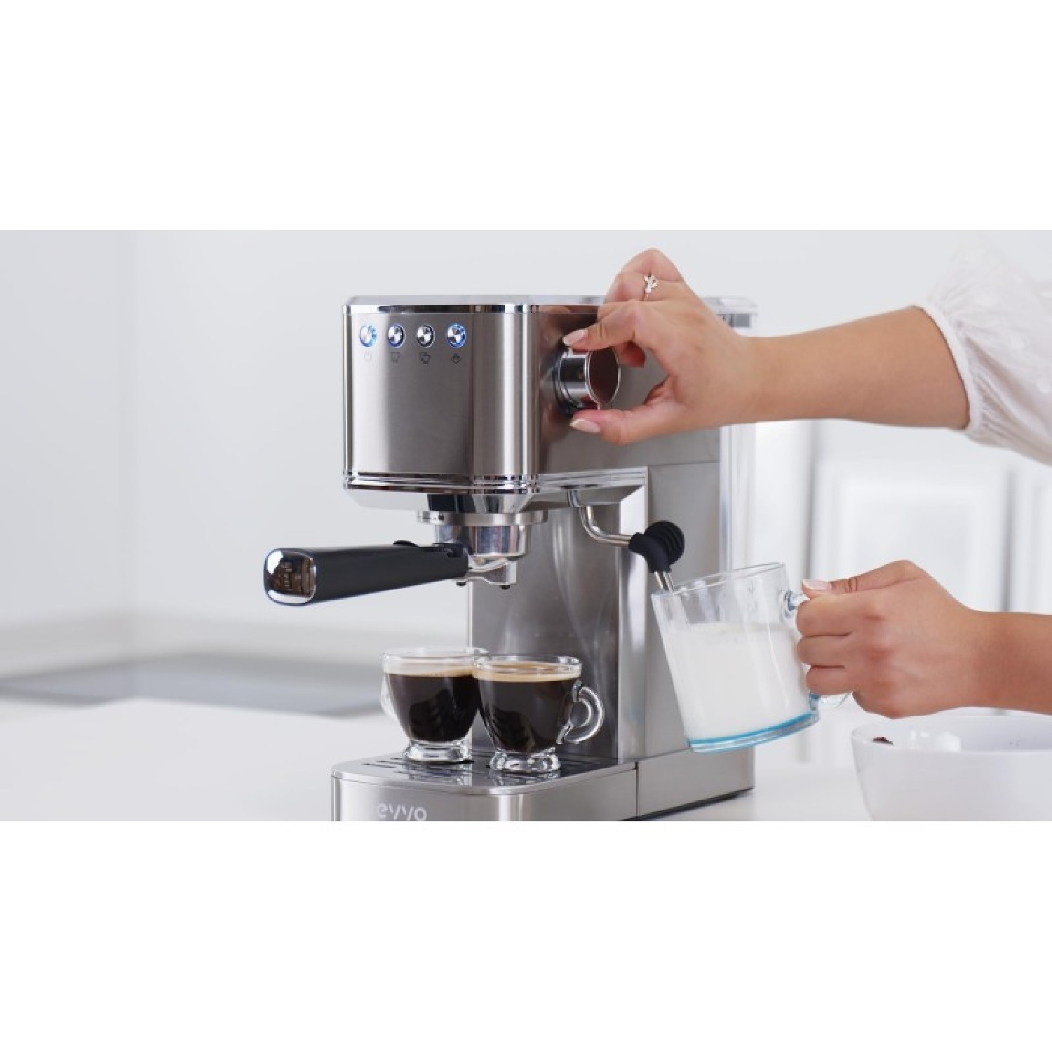 Una persona usa una cafetera plateada, girando una perilla mientras sostiene un vaso de leche.