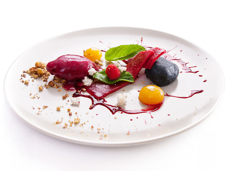 Postre gourmet con sorbetes de frutos rojos, bayas, menta, salsa y migas de nuez en un plato blanco.