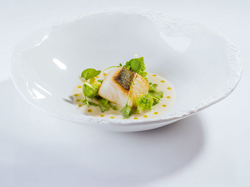 Un filete de pescado sellado en salsa clara con verduras verdes en un bol blanco hondo.