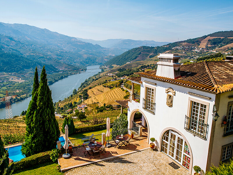 Una villa blanca con vistas a un valle, viñedos en terrazas y un río bajo un cielo azul.