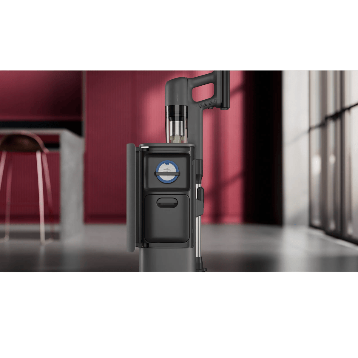 A black cordless vacuum cleaner stands upright with its accessory storage compartment open, set against a blurred red and grey room background.