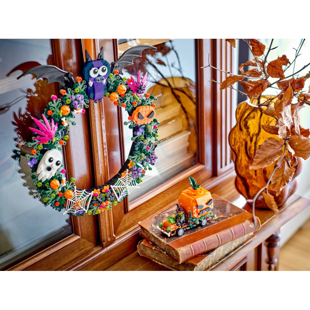 Wianek halloweenowy z klocków Lego wisi na drewnianej szafce obok książek i wazonu z liśćmi.