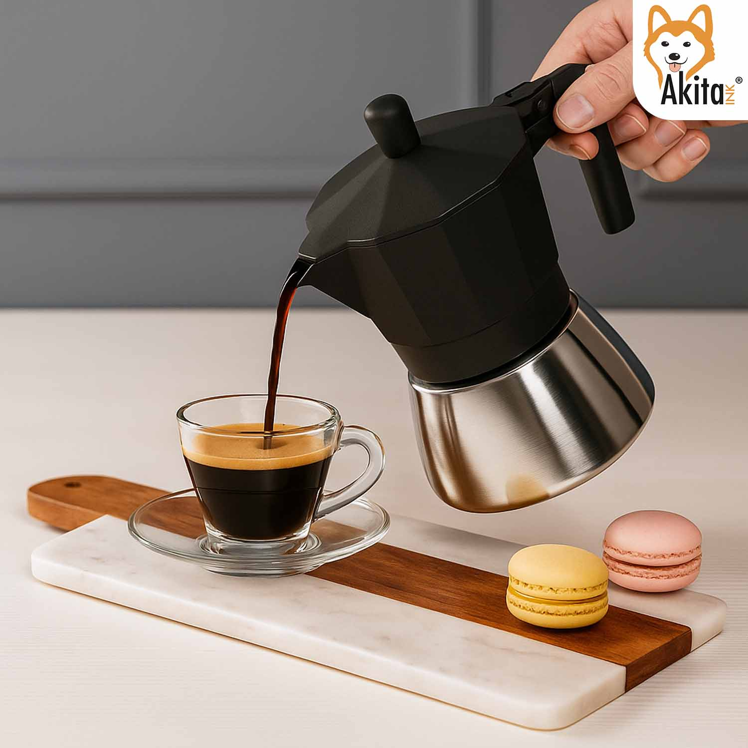 Una mano versa il caffè da una moka nera in una tazza di vetro su un tagliere di marmo e legno con macaron.