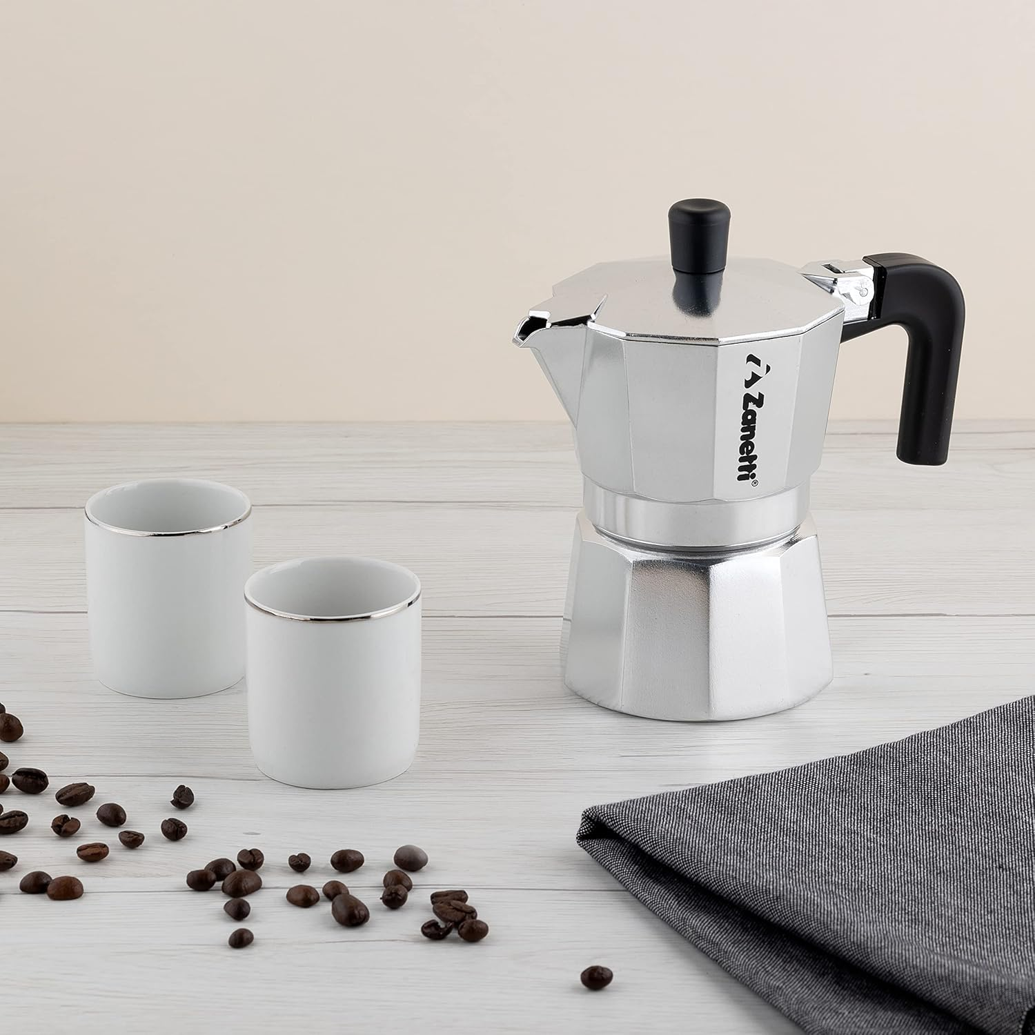 Una moka d'argento, due tazze bianche e chicchi di caffè su una superficie di legno bianco.