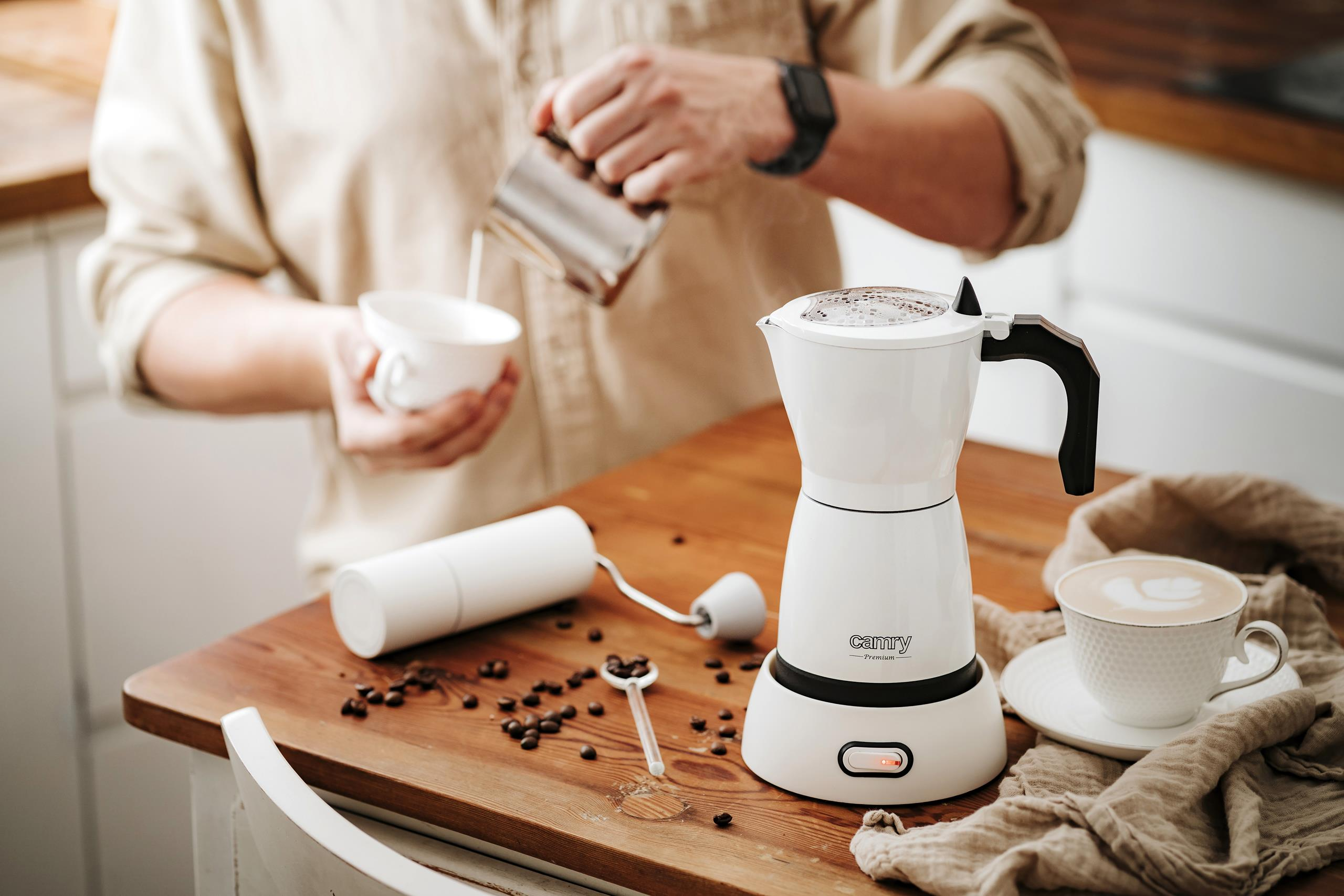 Una persona versa il latte in una tazza vicino a una moka bianca, un macinacaffè e chicchi di caffè.