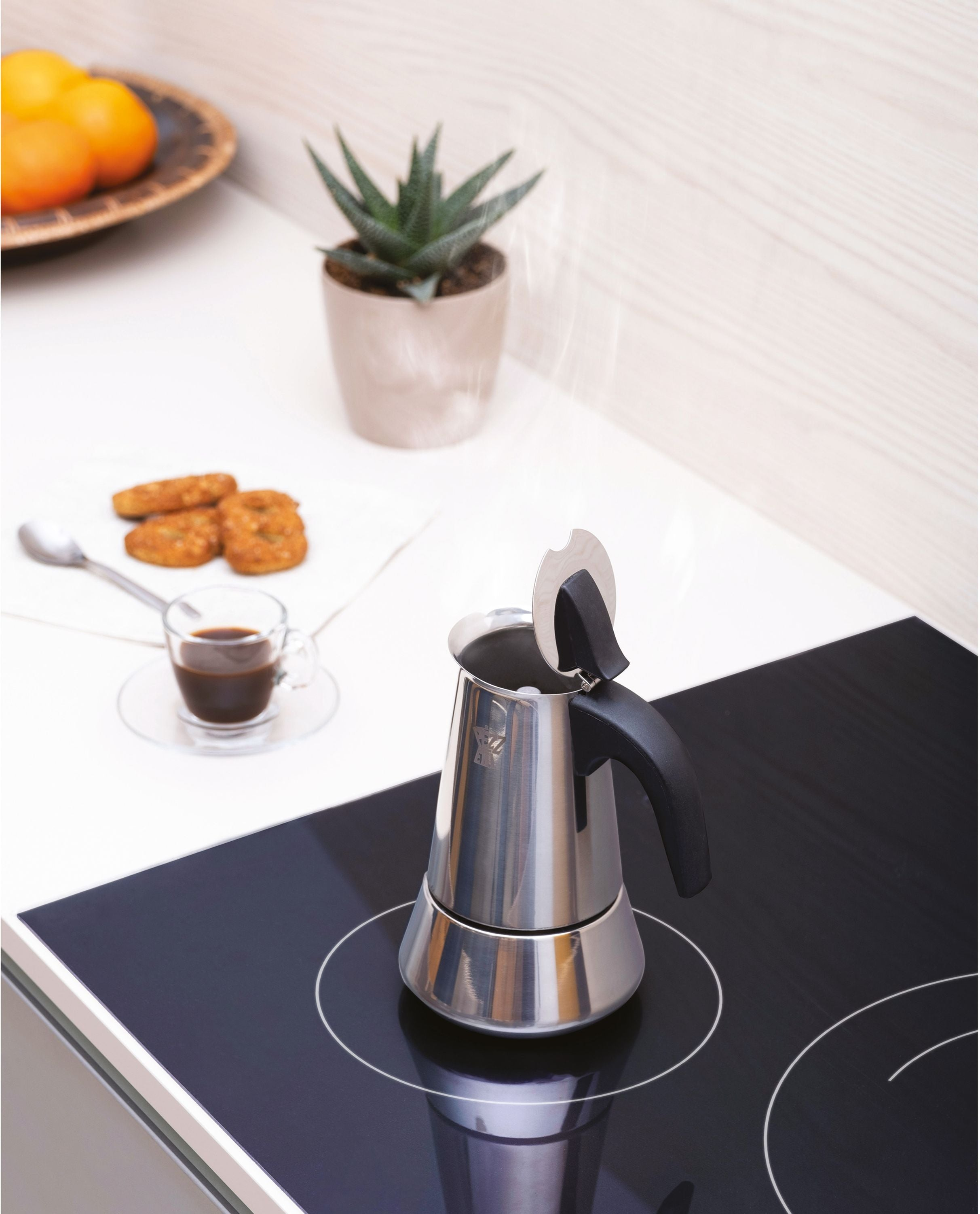 Una moka fumante su un piano cottura a induzione nero, con caffè, biscotti e una pianta vicini.