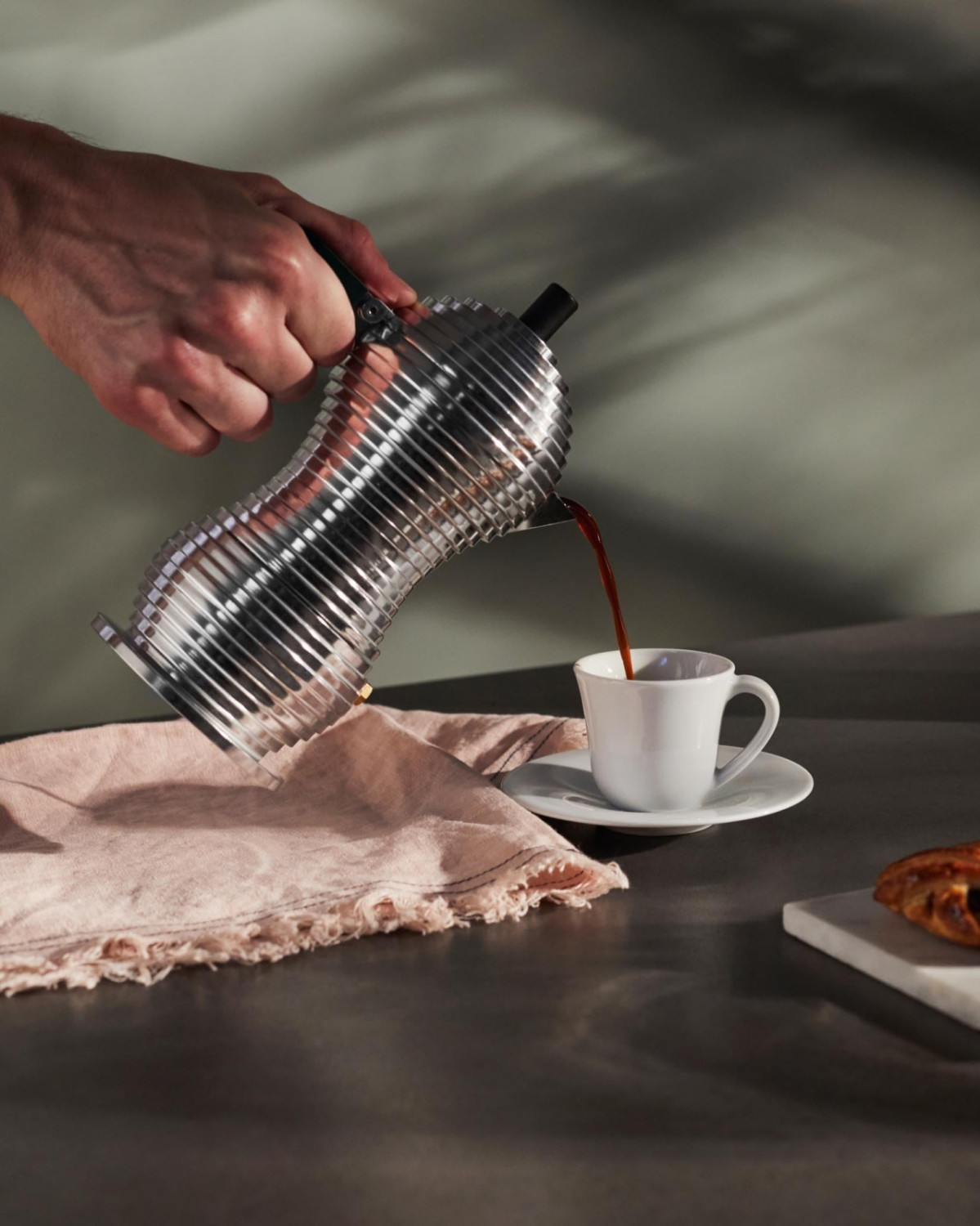 Una mano versa il caffè da una moderna caffettiera argentata in una tazza bianca su un tavolo.