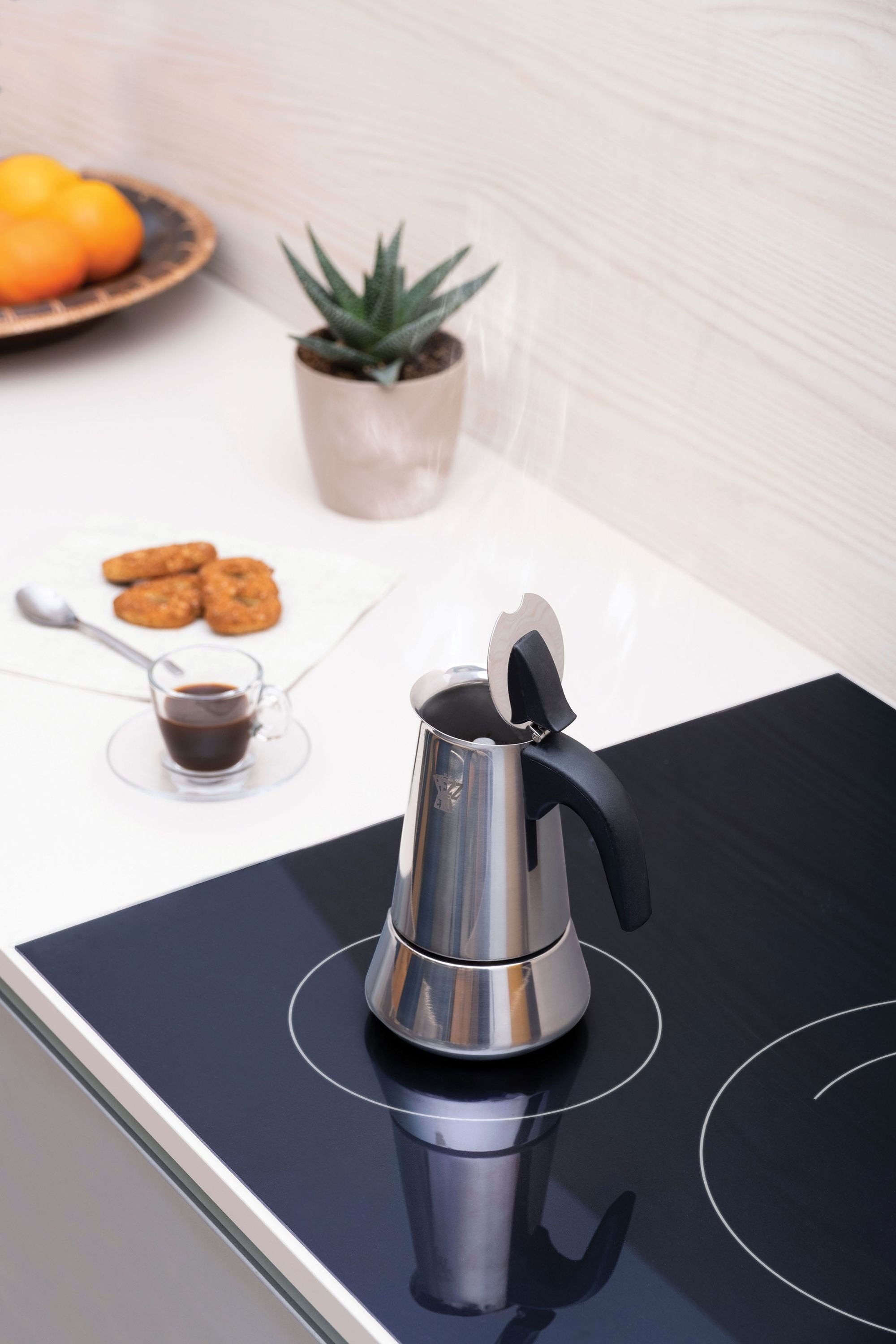 Una moka in acciaio inossidabile su un piano cottura a induzione nero, con espresso, biscotti, una pianta e arance.