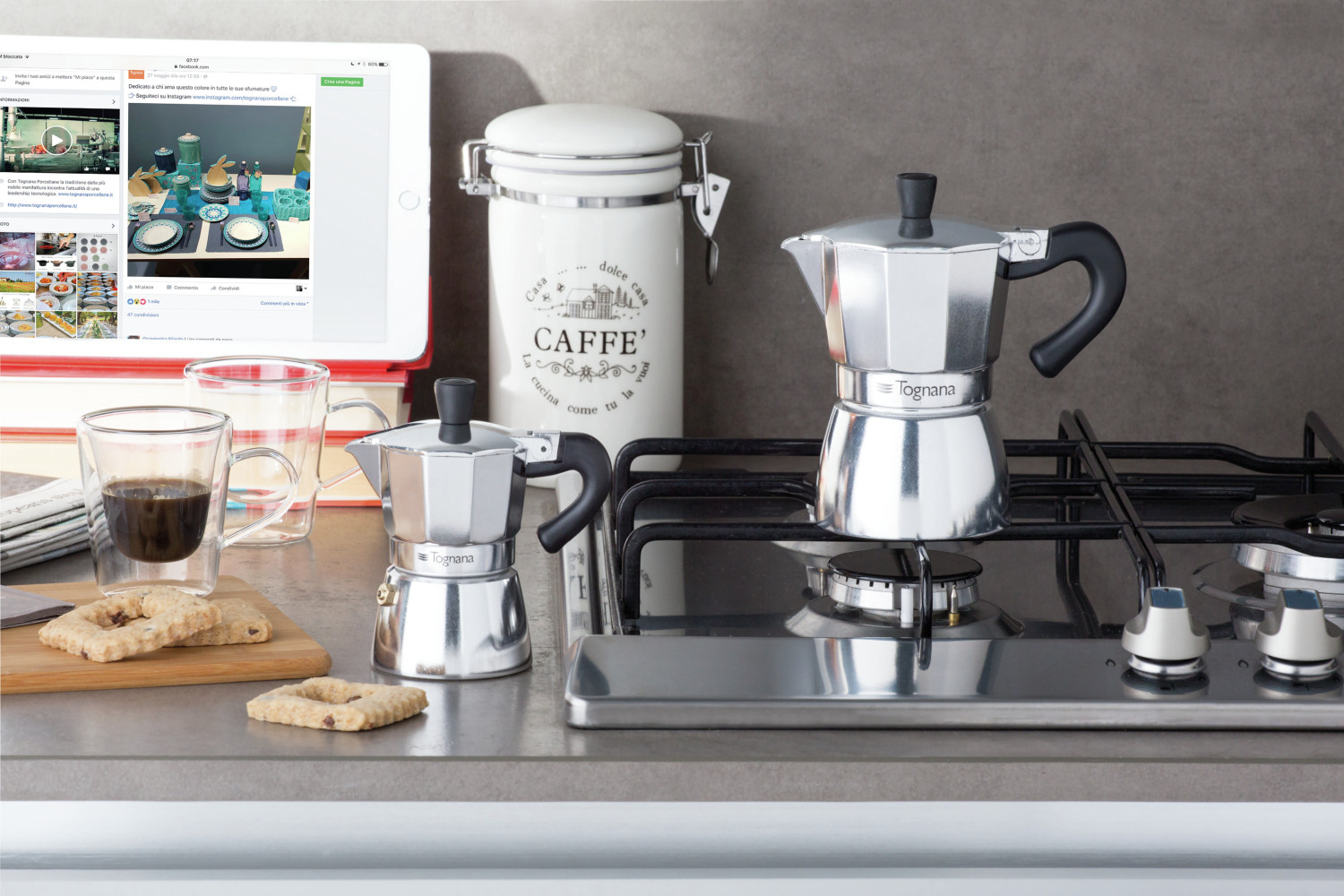 Due caffettiere, tazze da caffè e un barattolo si trovano su un piano cucina vicino a un fornello e a un tablet.