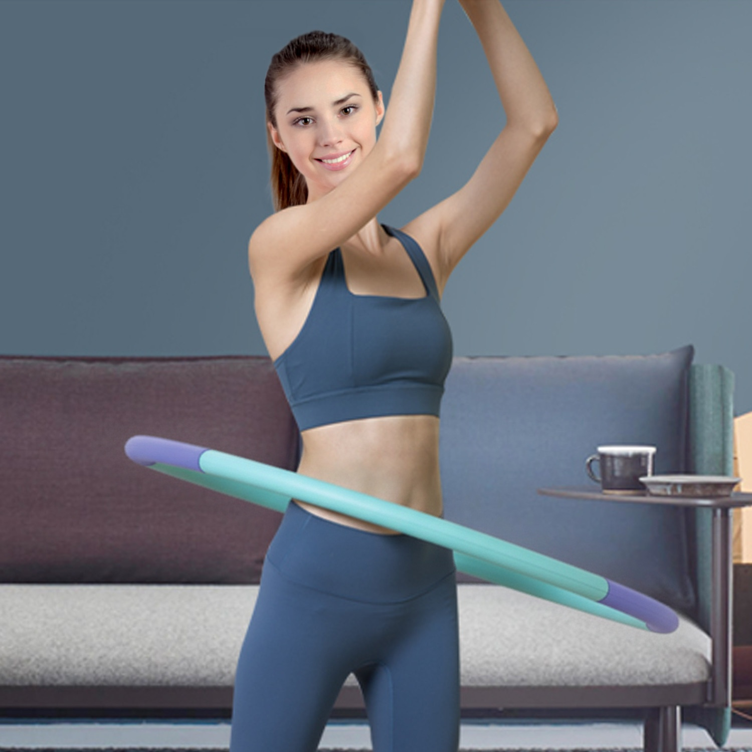 Eine Frau in blauer Sportkleidung lächelt, während sie einen blauen Hula-Hoop-Reifen im Wohnzimmer nutzt.