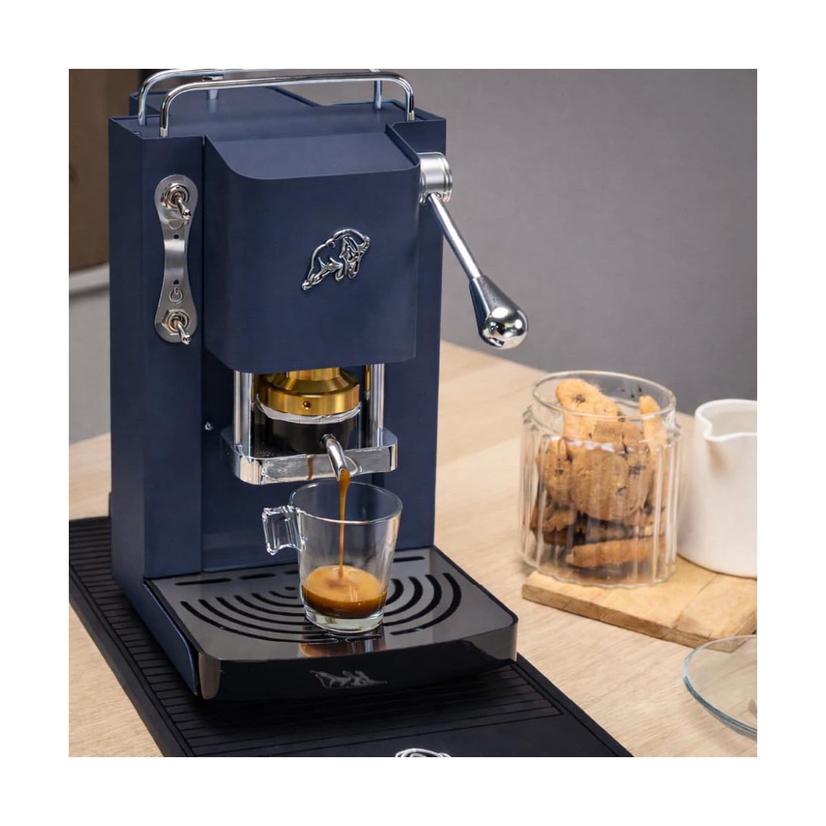 Eine blaue Espressomaschine brüht Kaffee in eine Glastasse auf einem Holztisch neben Keksen.