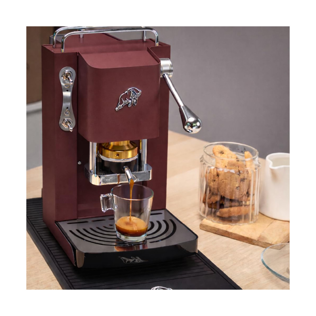 Eine bordeauxrote Espressomaschine brüht Kaffee in eine Glastasse. Daneben stehen Kekse in einem Glas.