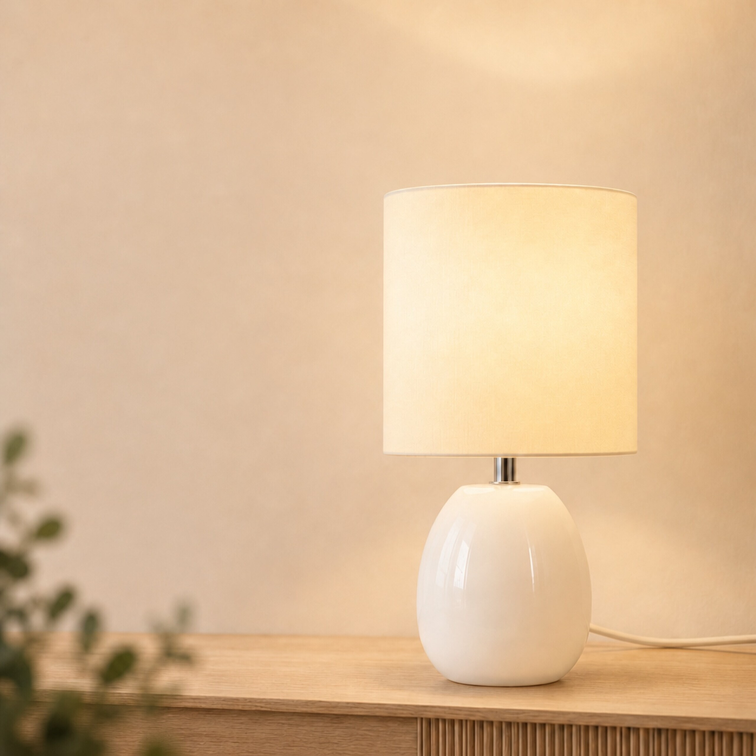 Eine weiße Tischlampe mit zylindrischem Schirm steht auf einer Holzkommode vor einer beigen Wand.