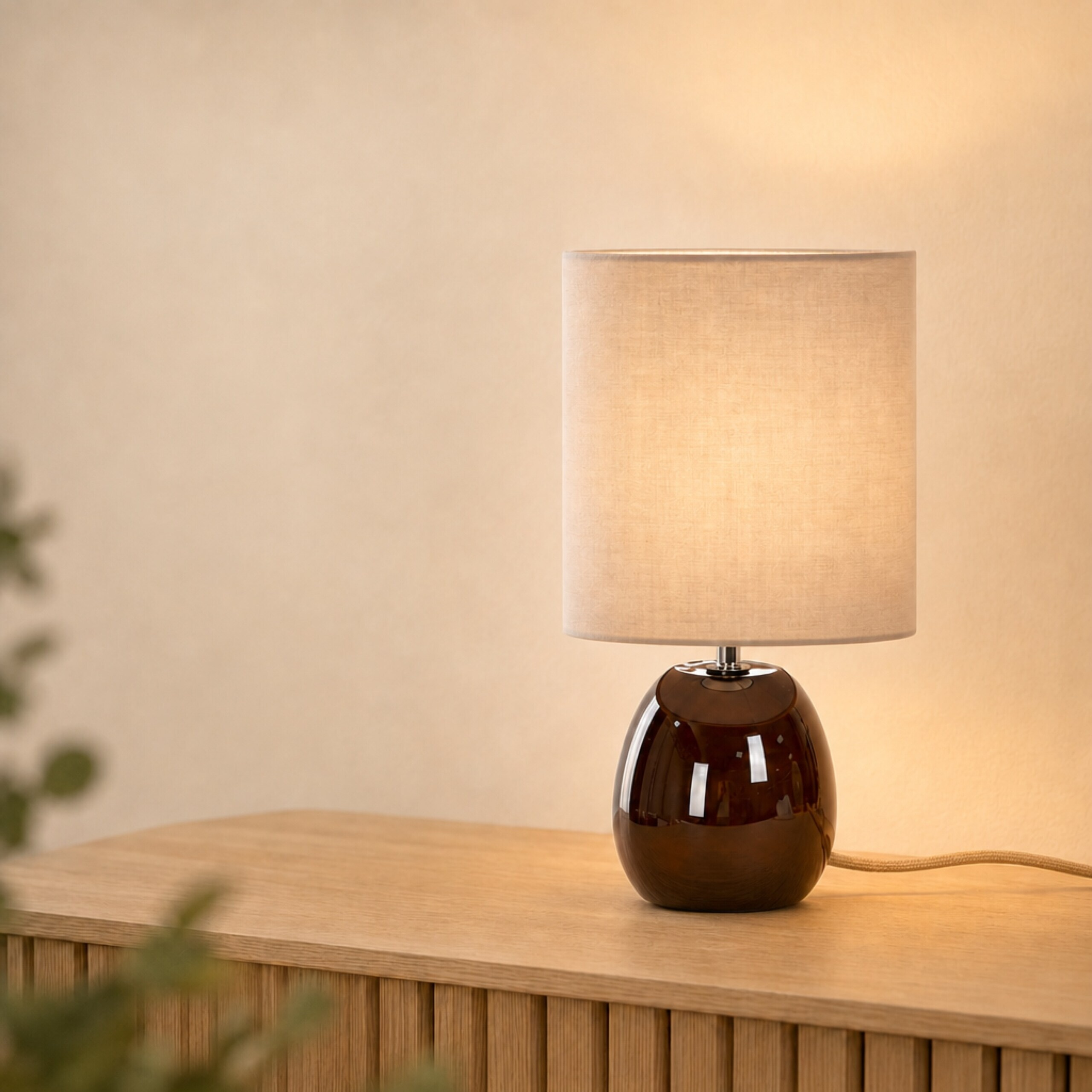 Eine kleine Tischlampe mit dunklem Keramikfuß und beigem Schirm steht auf einer Holzkommode.