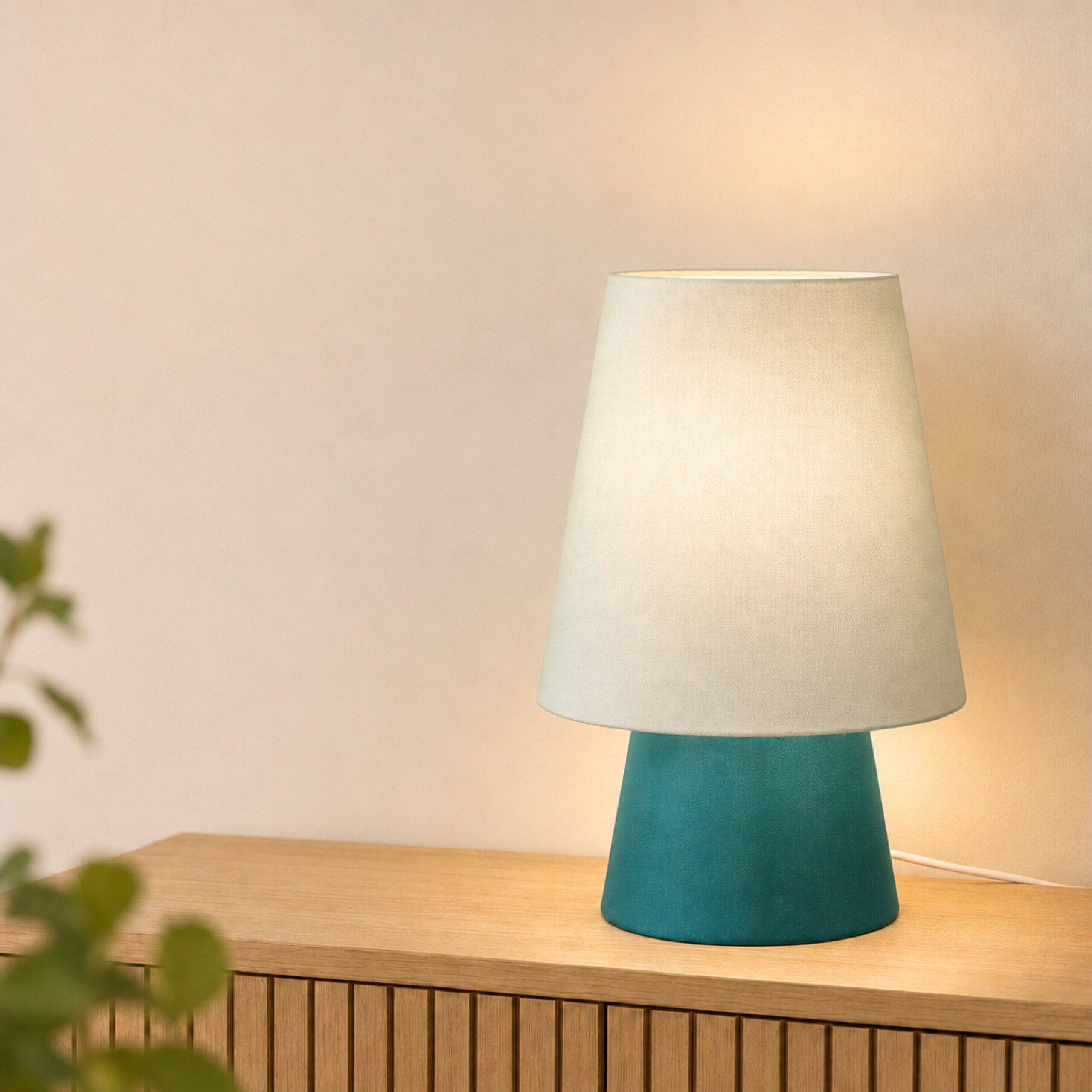 Eine blau-türkise Tischlampe mit hellem Lampenschirm steht auf einem Holzschrank vor einer beigen Wand.