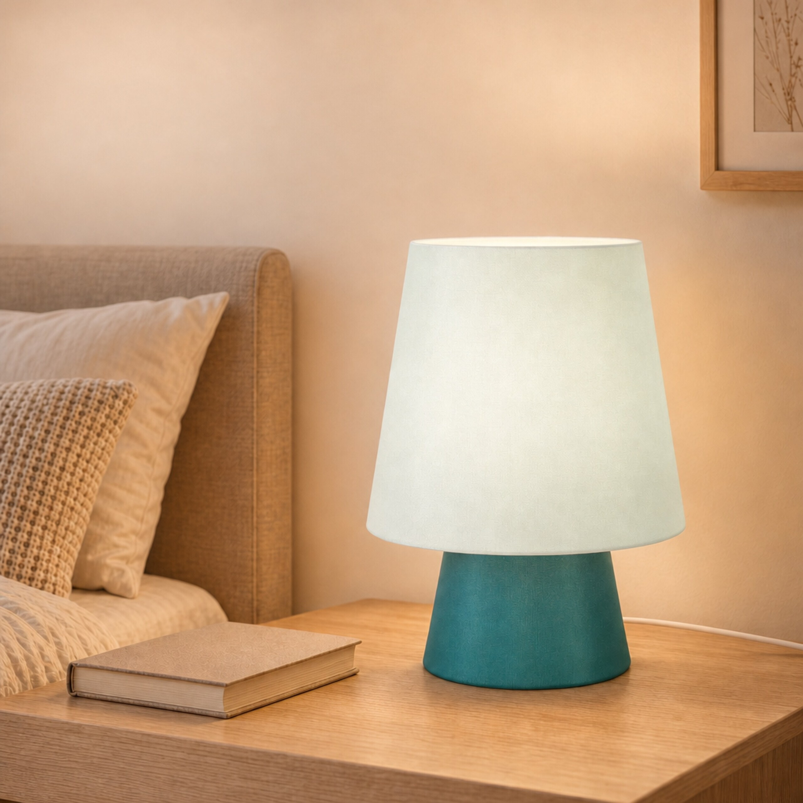 Eine erleuchtete Tischlampe mit blauem Sockel steht neben einem Bett auf einem Holztisch, daneben ein Buch.