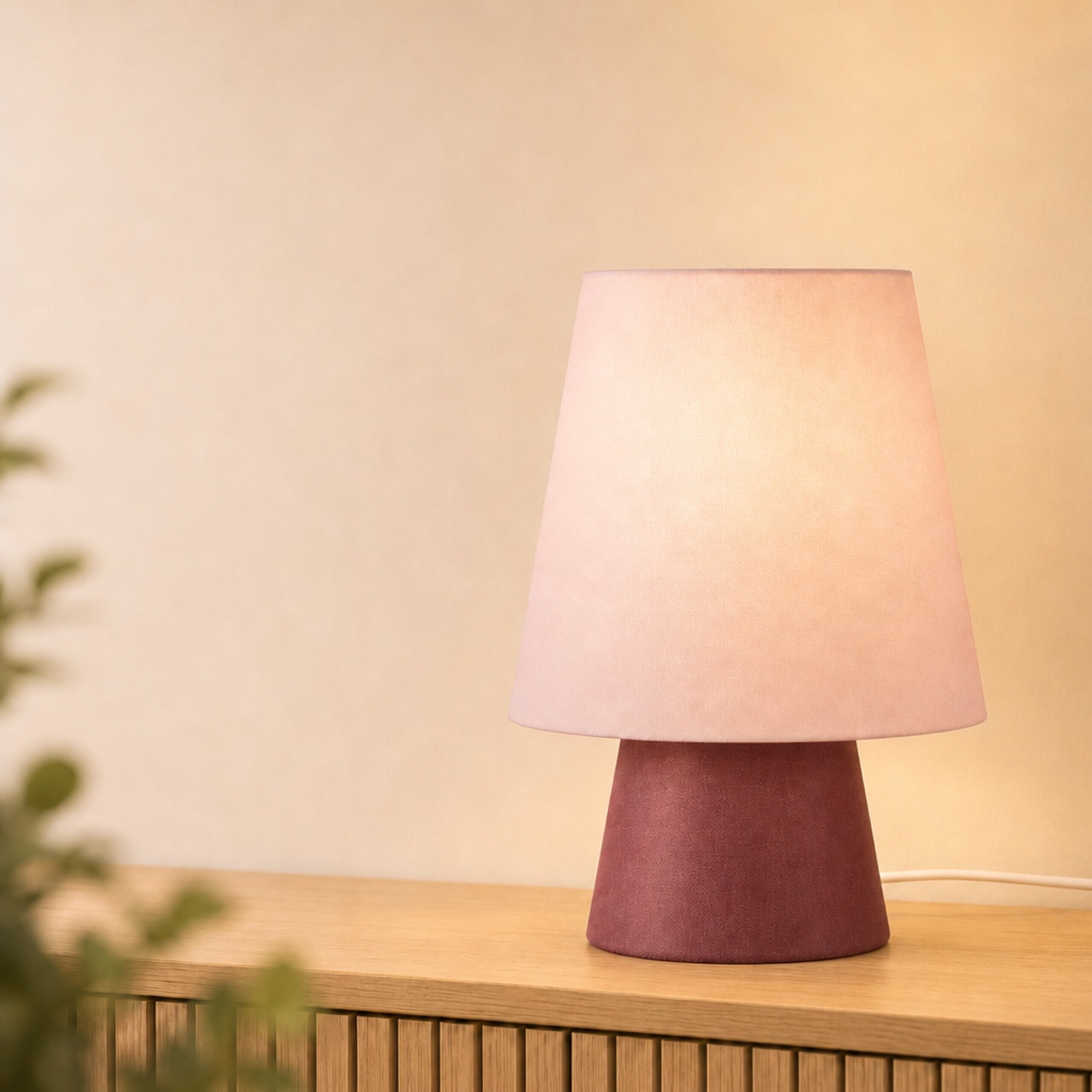 Eine rosafarbene Tischlampe mit kegelförmigem Schirm steht auf einer Holzkommode vor einer beigen Wand.