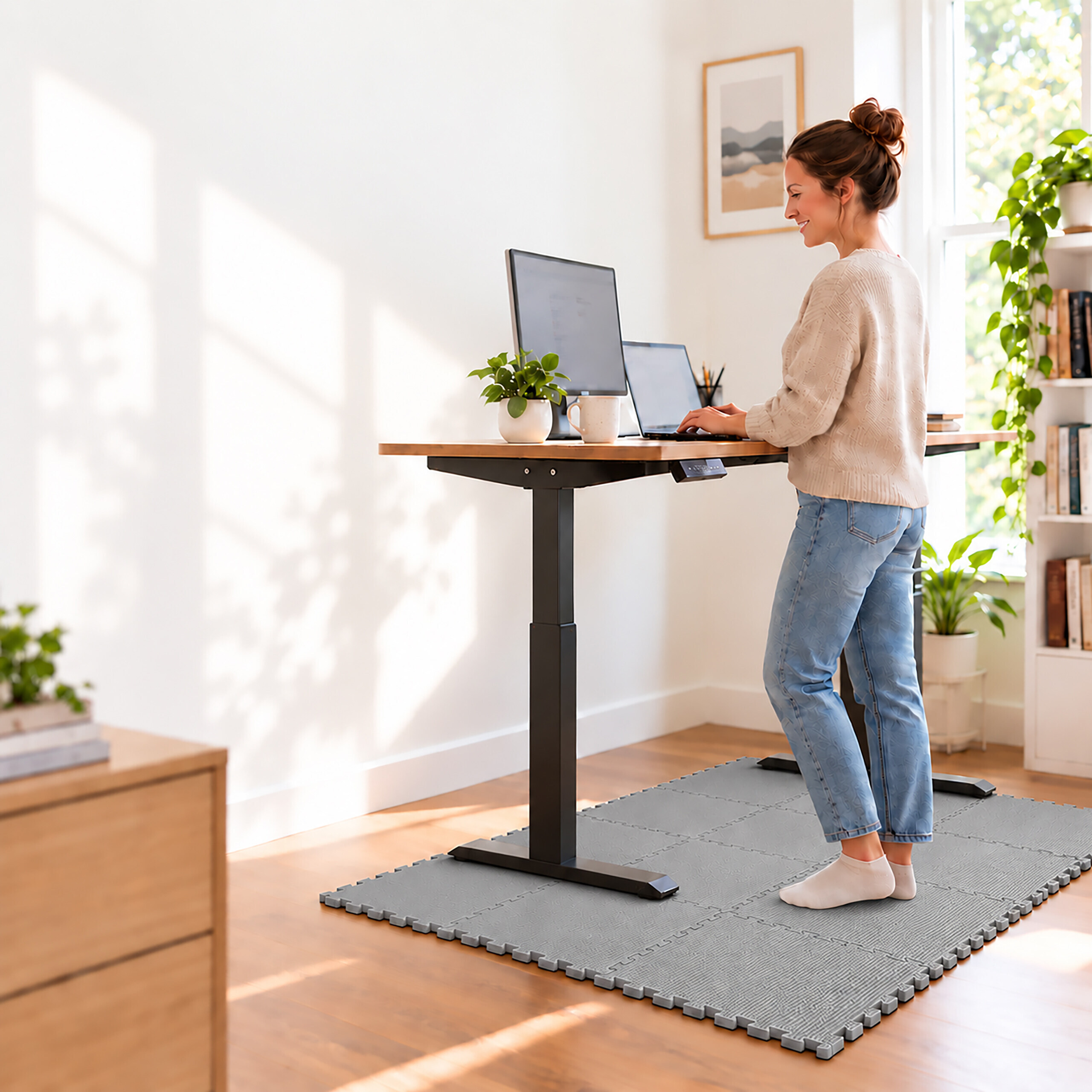 Frau arbeitet im stehenden Zustand an einem höhenverstellbaren Holztisch mit Monitor und Laptop, auf einer grauen Matte.