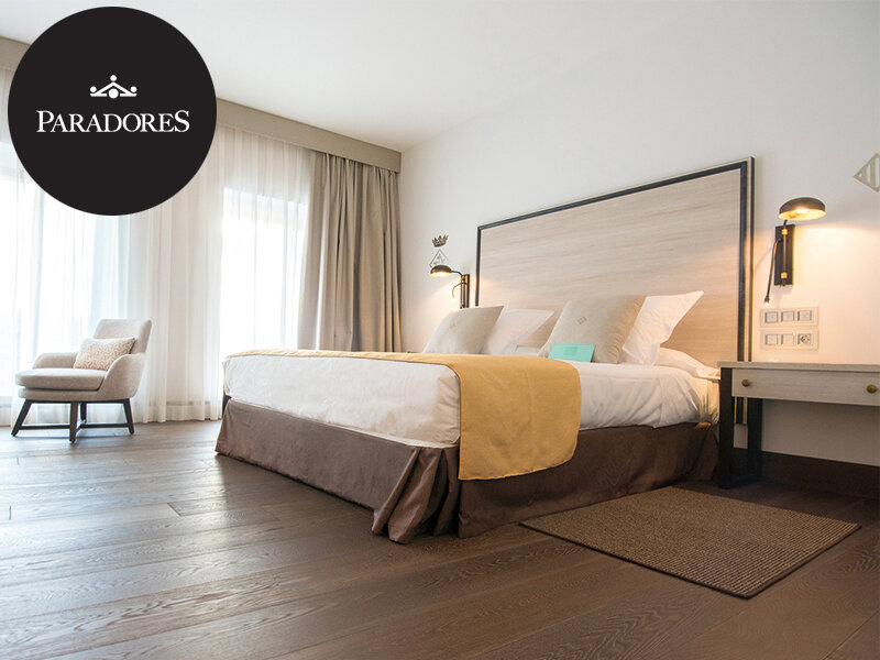 Habitación de hotel moderna con cama grande, suelo de madera, sillón y grandes cortinas.