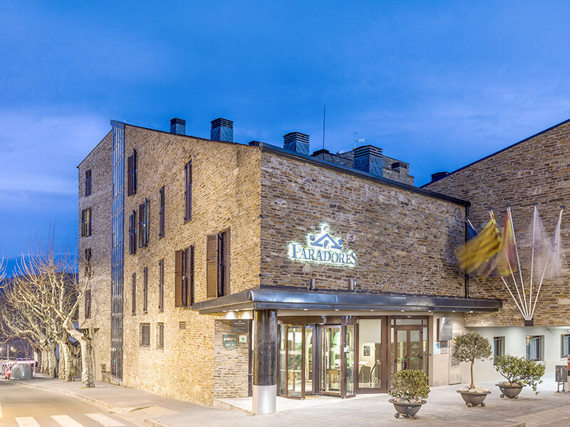 Un hotel Paradores iluminado con fachada de piedra, entrada de cristal y banderas ondeando al anochecer.