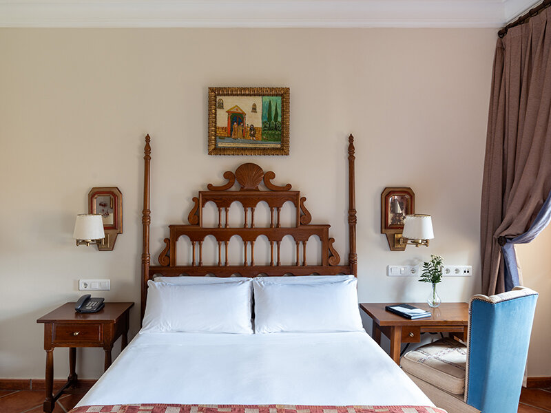 Una acogedora habitación de hotel con una cama de madera grande, almohadas blancas, mesitas de noche y una silla azul.