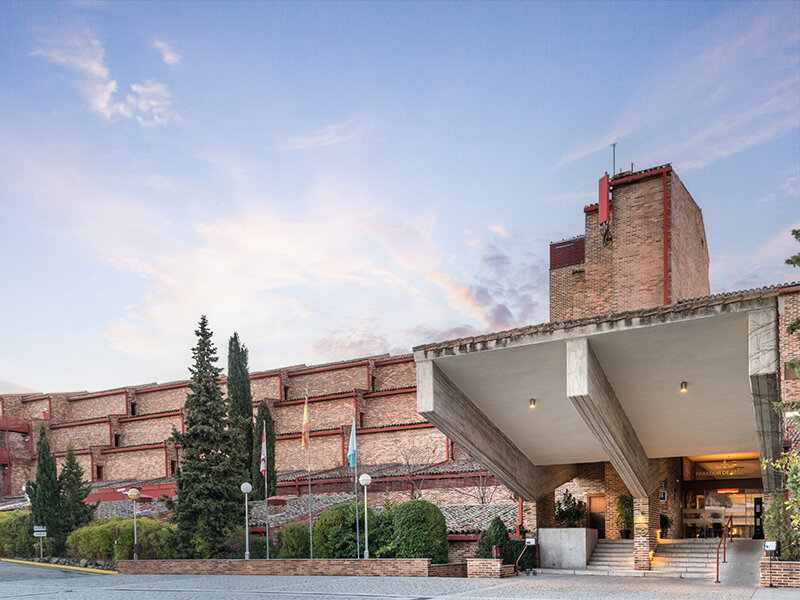 Exterior de hotel moderno de ladrillo con gran marquesina de hormigón y árboles al atardecer.