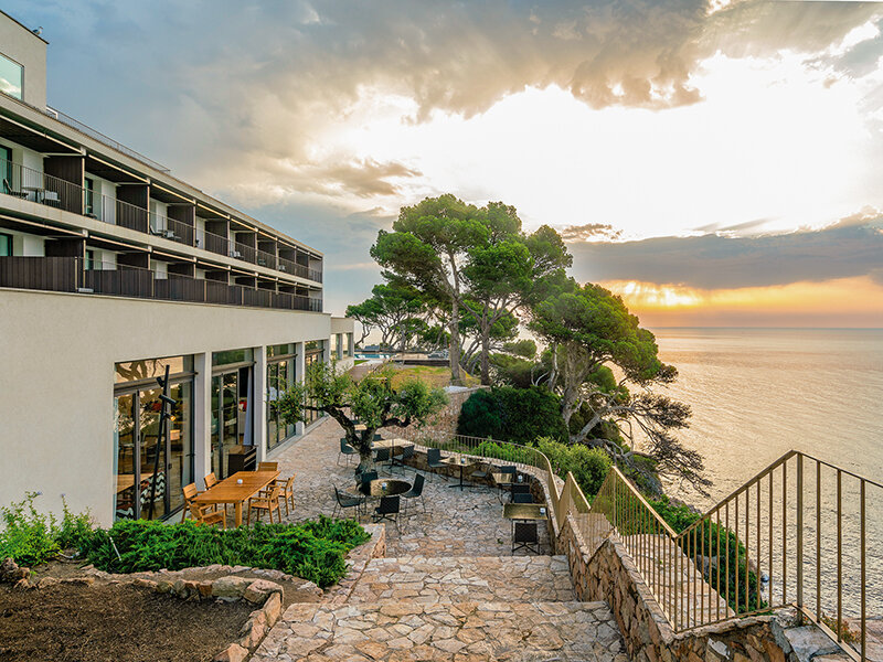 Terraza de hotel costero con mesas, árboles, escaleras de piedra y vista al atardecer sobre el mar.