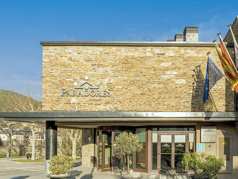 Exterior del hotel Paradores con fachada de piedra, entrada de vidrio giratoria, plantas en maceta y banderas bajo un cielo azul.