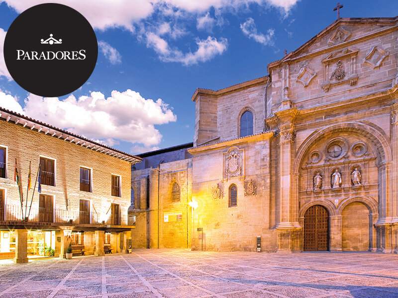Edificios históricos de piedra y una iglesia en una plaza iluminada bajo un cielo azul con nubes.